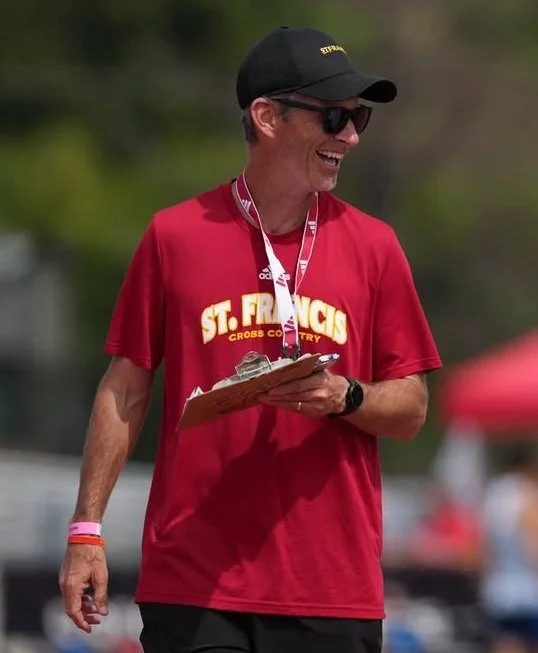 A man wearing a red 'St. Francis Cross Country' T-shirt, a black cap, sunglasses, and a wristwatch, smiling while holding a clipboard and pen at an outdoor event.