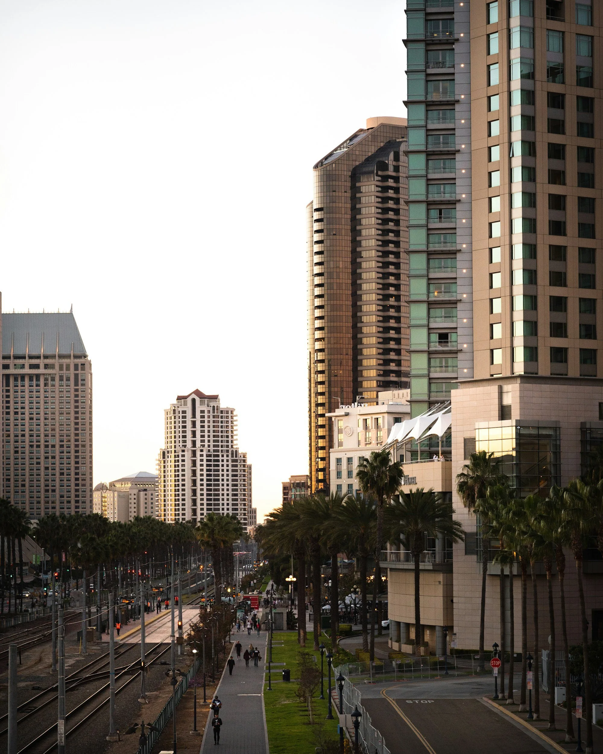 Cityscape with tall commercial buildings, palm trees, and a train station platform in the foreground. Pedestrians are walking along the sidewalk, and the scene is lit by fading daylight.