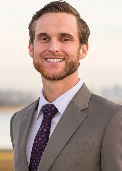 Portrait of a smiling young man in a gray suit, light purple shirt, and purple patterned tie outdoors with a blurred cityscape background.