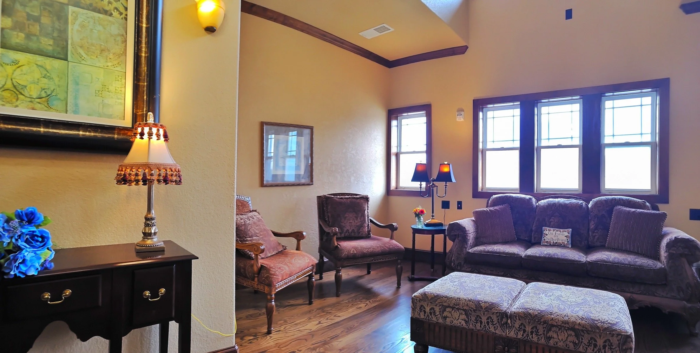 Living room with yellow walls, hardwood floors, large windows, and vintage furniture including patterned armchairs, a floral sofa, and side tables with lamps.