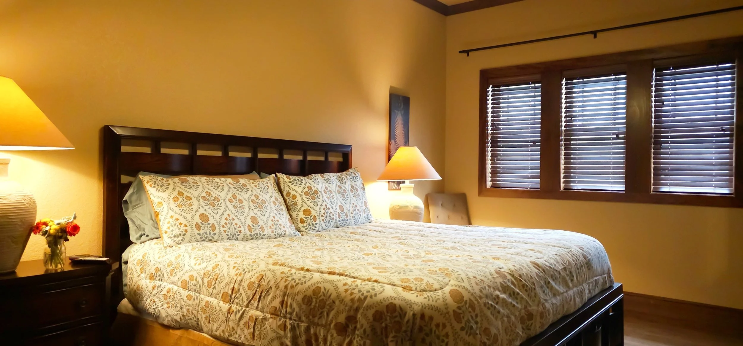 A cozy bedroom with a large bed featuring floral-patterned bedding and pillows, two bedside tables each with a lamp, a small chair, and three window blinds on the window.