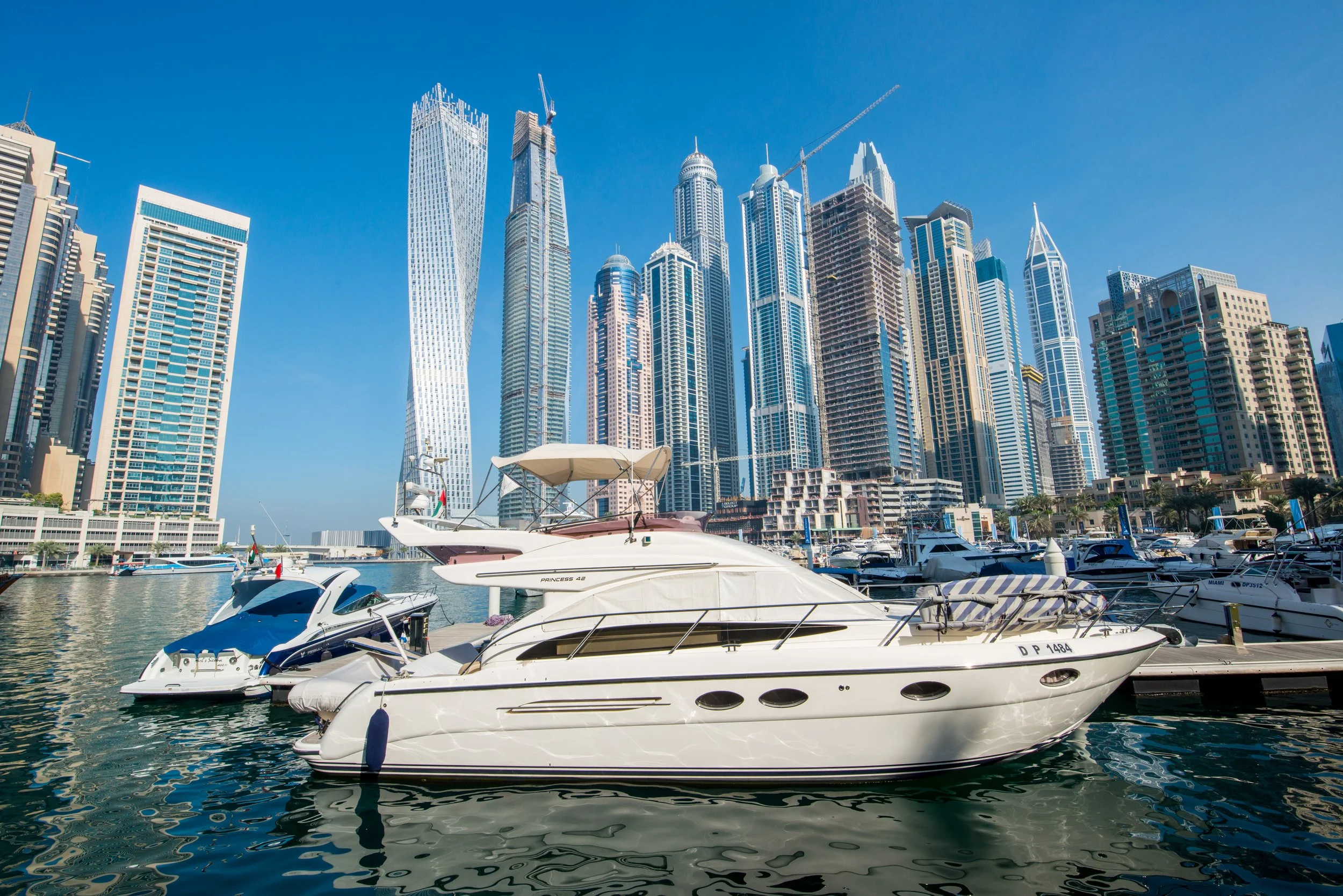 A marina with several white yachts docked in calm water, with high-rise buildings and construction cranes visible in the background under a clear blue sky.