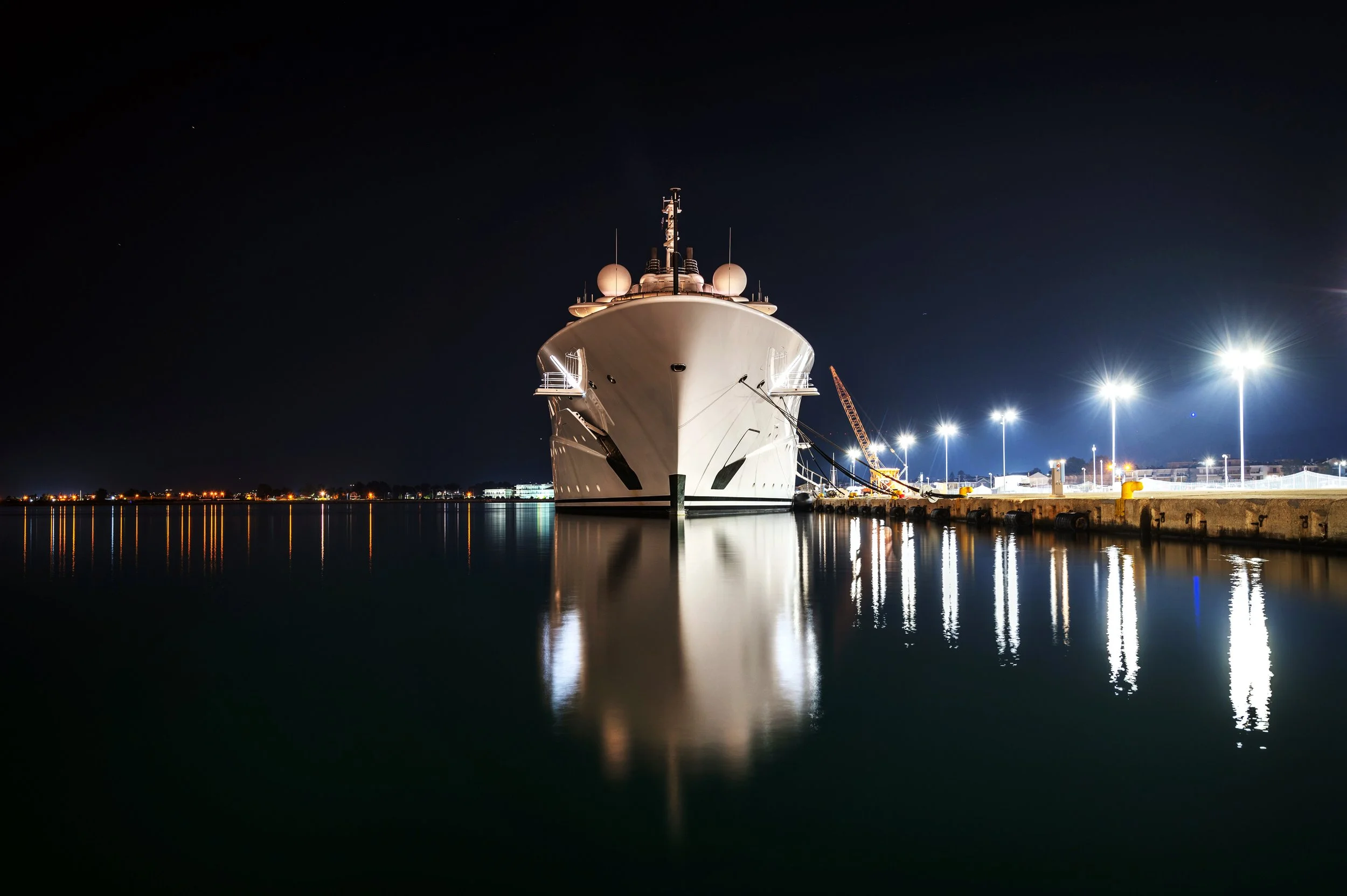 A large white yacht docked at a marina at night, illuminated by bright lights with reflections on the water, and a dark starry sky above.