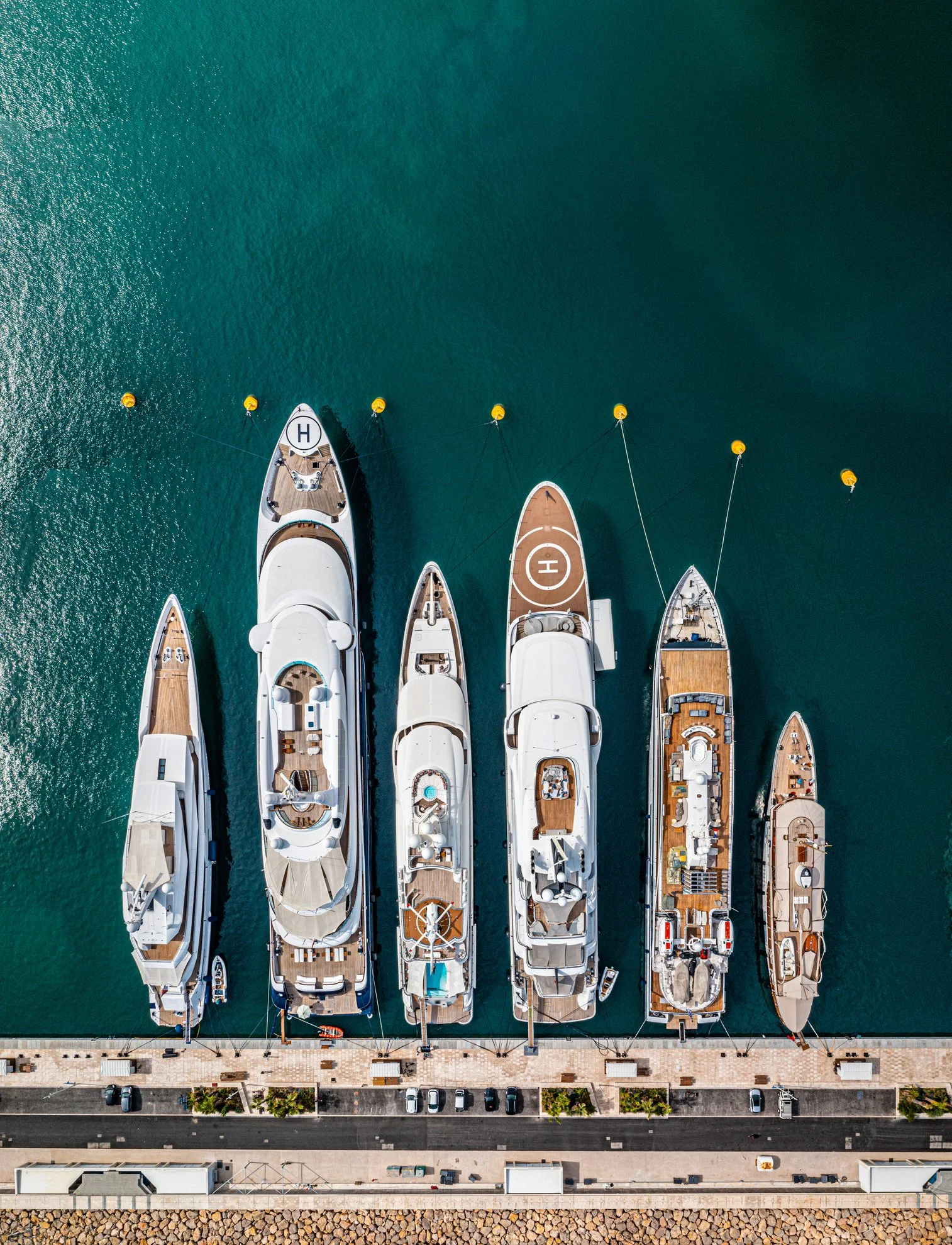 Aerial view of yachts docked at a marina, with several boats lined up side by side and a sidewalk with parked cars below.