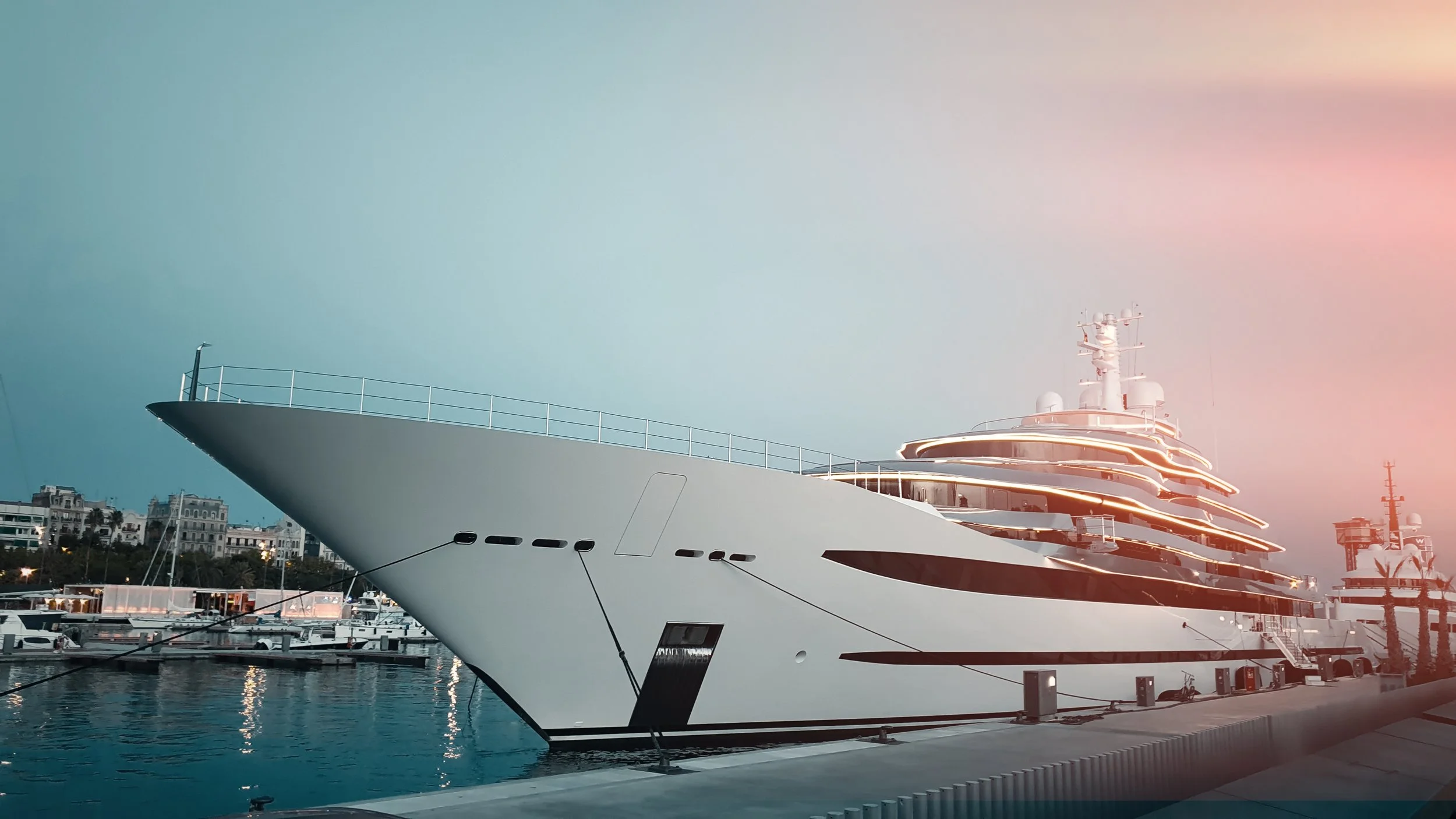Large luxury yacht docked at marina with city buildings in the background during sunset, featuring sleek white exterior and illuminated decks.