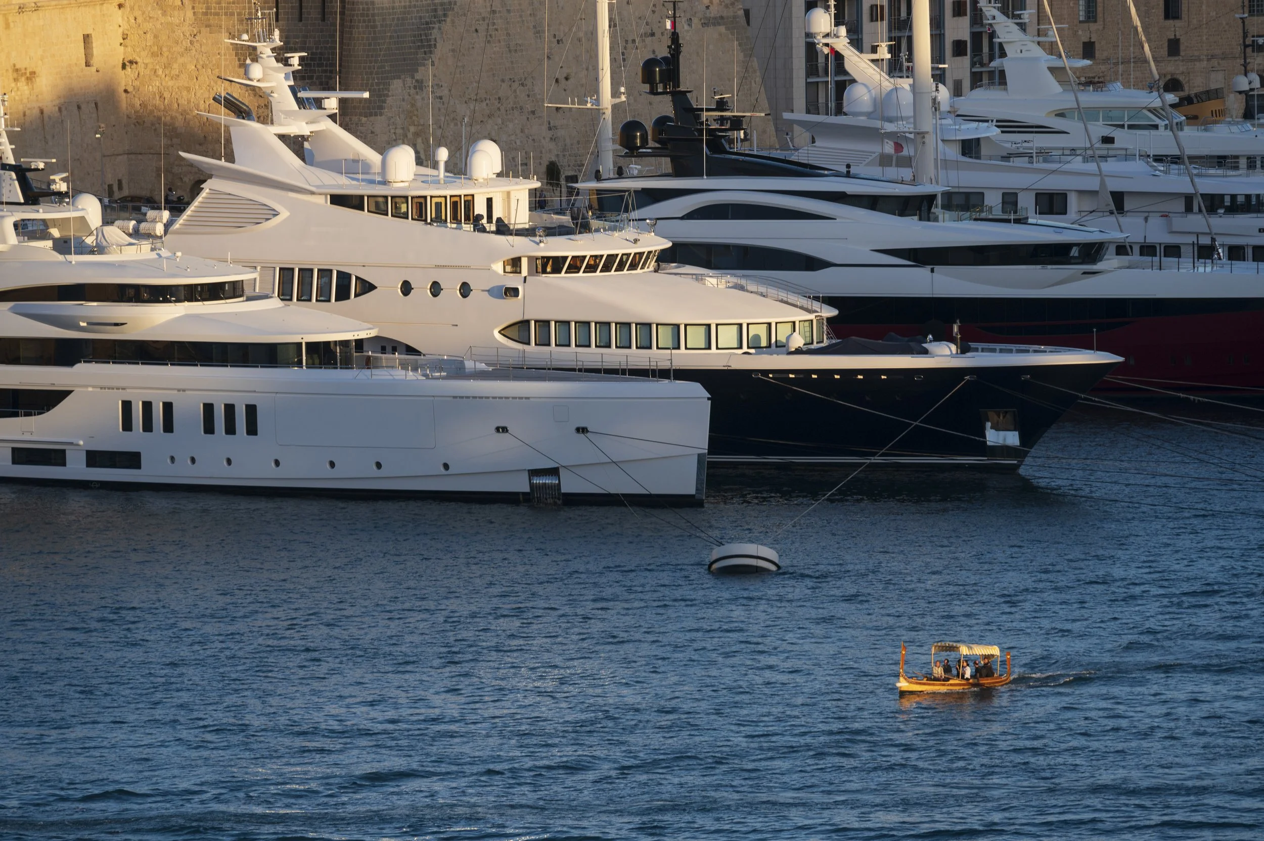 Several large yachts docked along a stone wall, with a small wooden boat with six people in the water nearby.