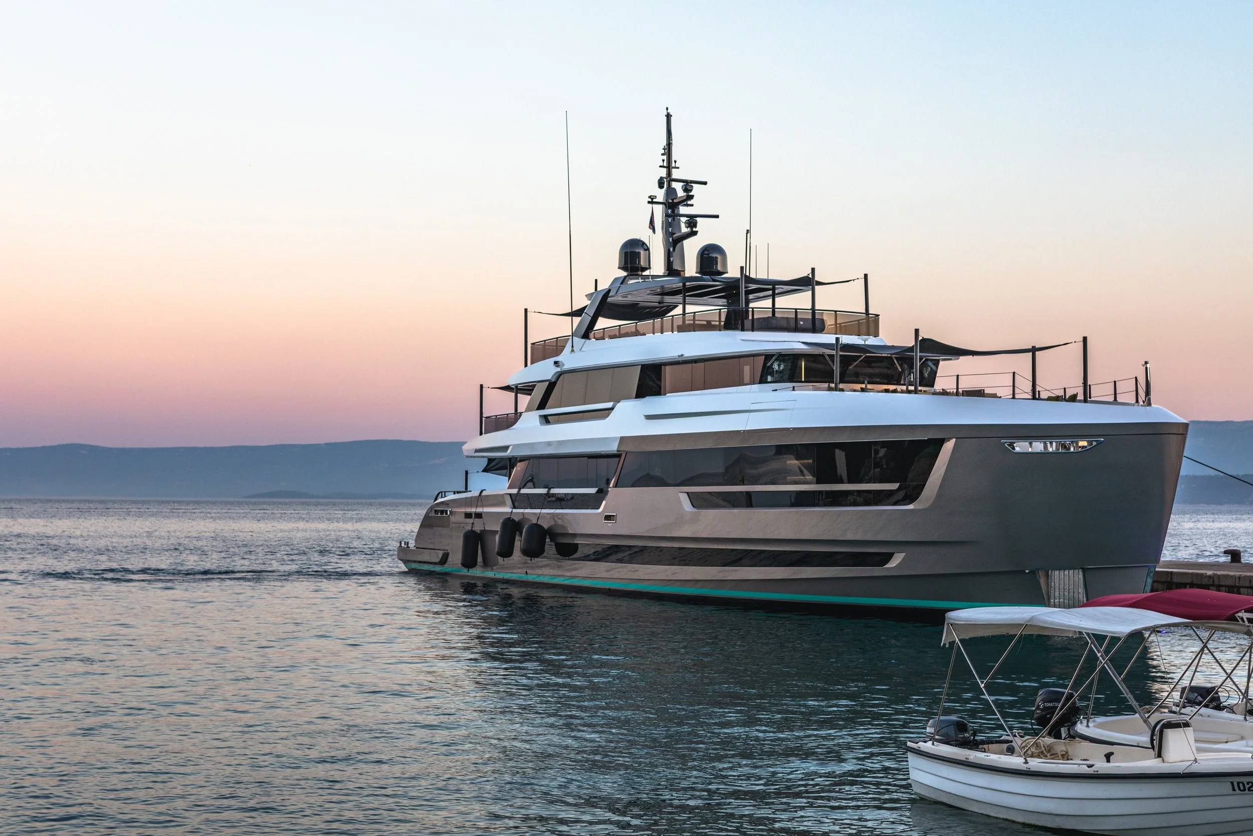A large white yacht docked in a harbor during sunset with a smaller boat nearby and calm water in the background.