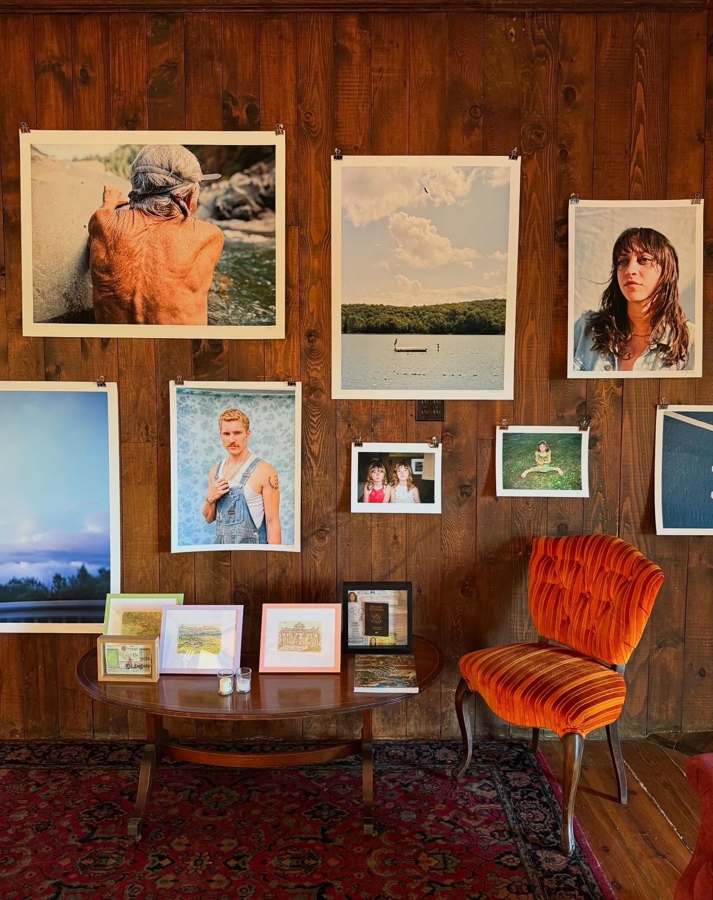 A wood-paneled wall decorated with various framed photographs of people and landscapes, a small table with framed artwork and candles, and an orange striped upholstered chair.