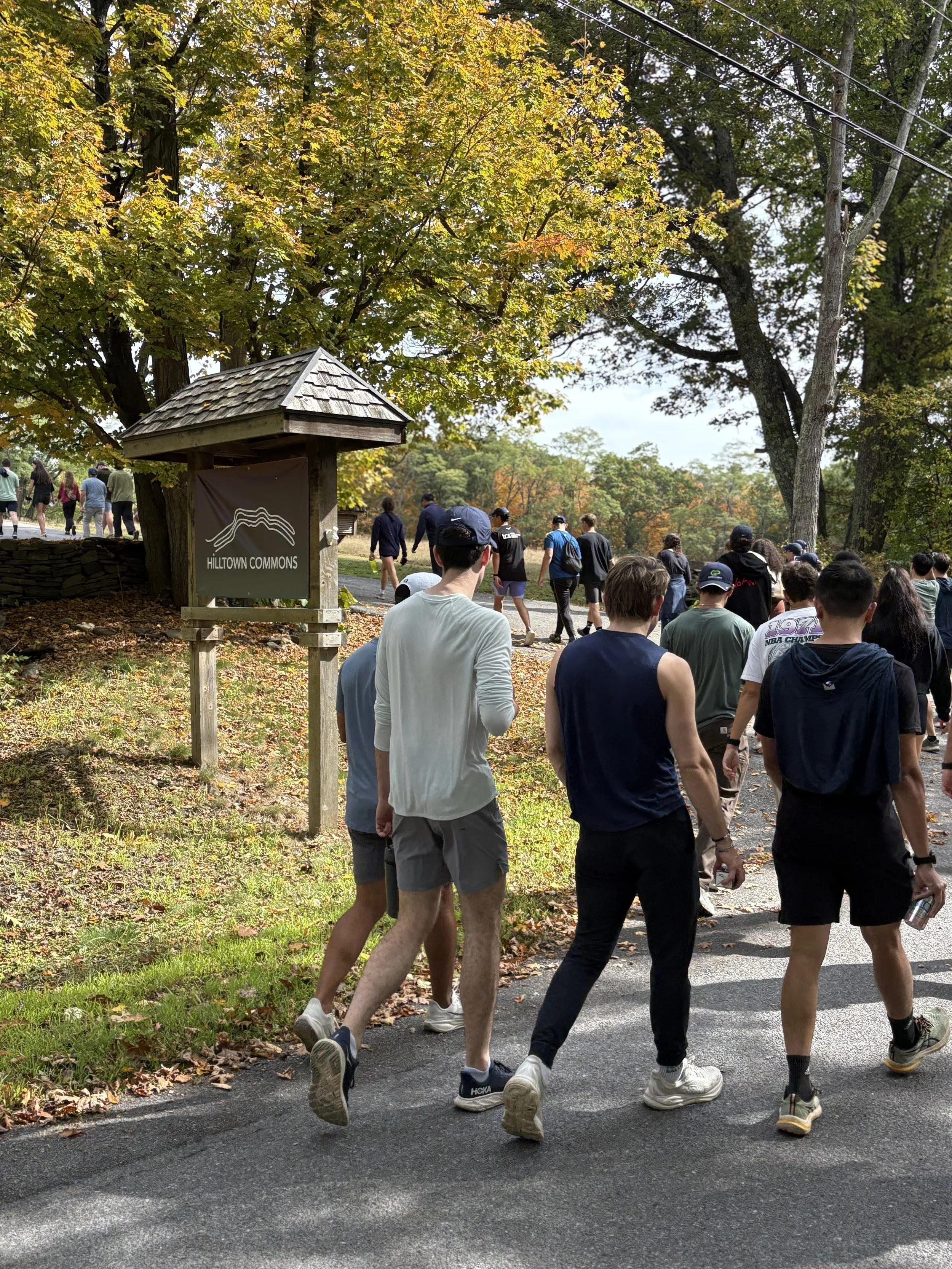 People walking and jogging in a park during autumn, with trees showing fall foliage and a sign reading 'HILLTOWN COMMONS' near the path.
