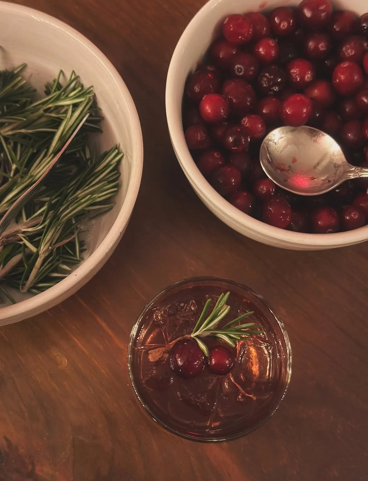 Bowl of fresh cranberries, bowl of rosemary sprigs, glass of cranberry cocktail with rosemary garnish on a wooden table.