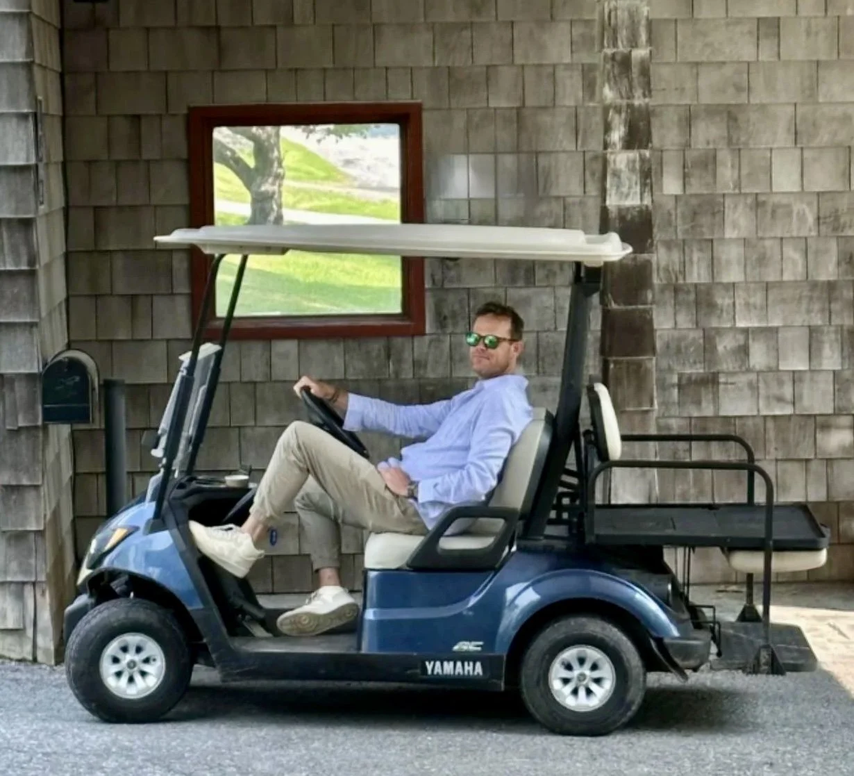 Man sitting on a blue golf cart, wearing sunglasses, a light blue shirt, and beige pants, parked in front of a rustic building with shingle siding and a window showing a green landscape.