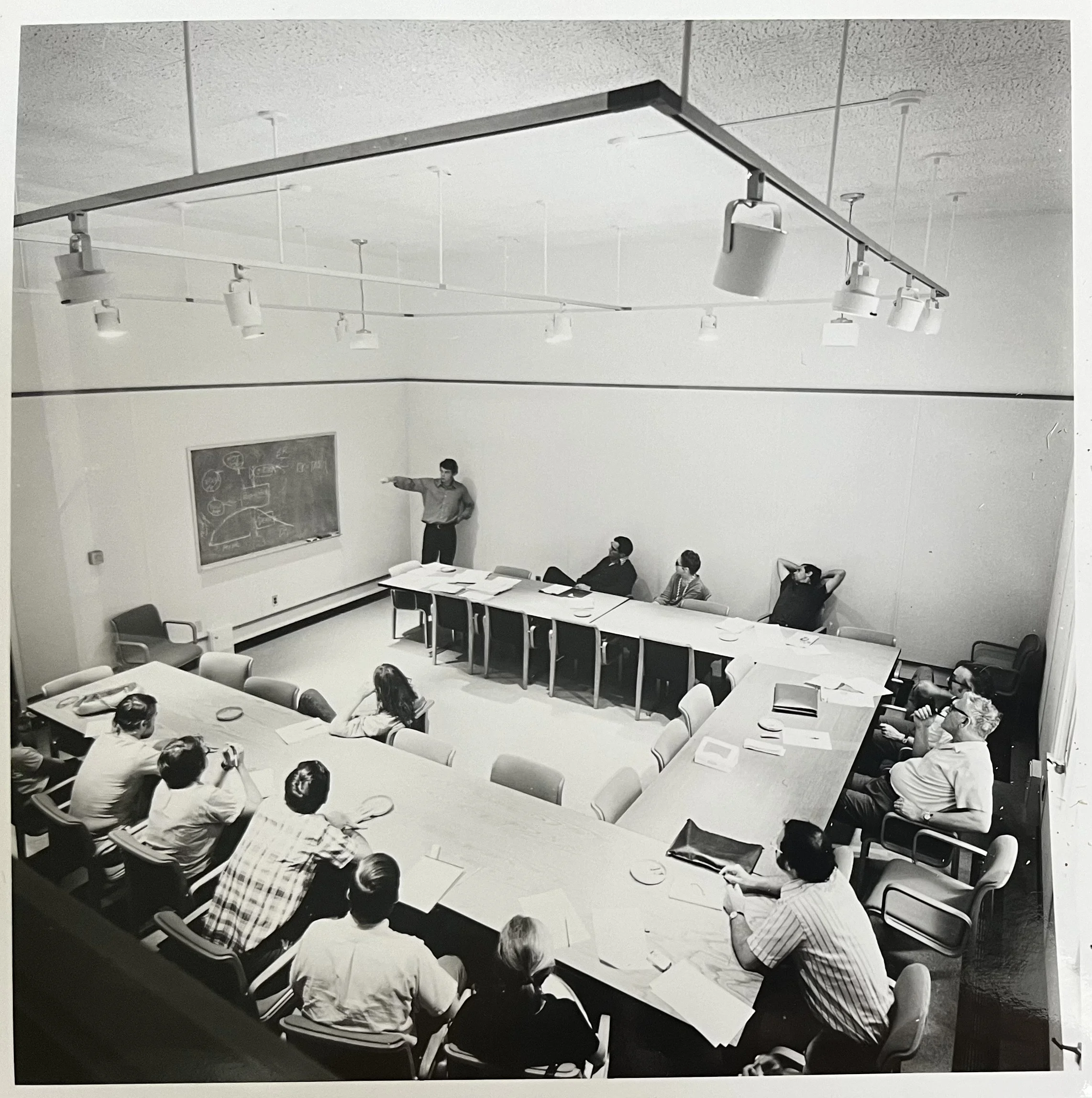 A black and white photo of a classroom or conference room with a teacher or presenter standing at the blackboard, pointing at it, while several adults are seated around a large U-shaped table, listening or taking notes.
