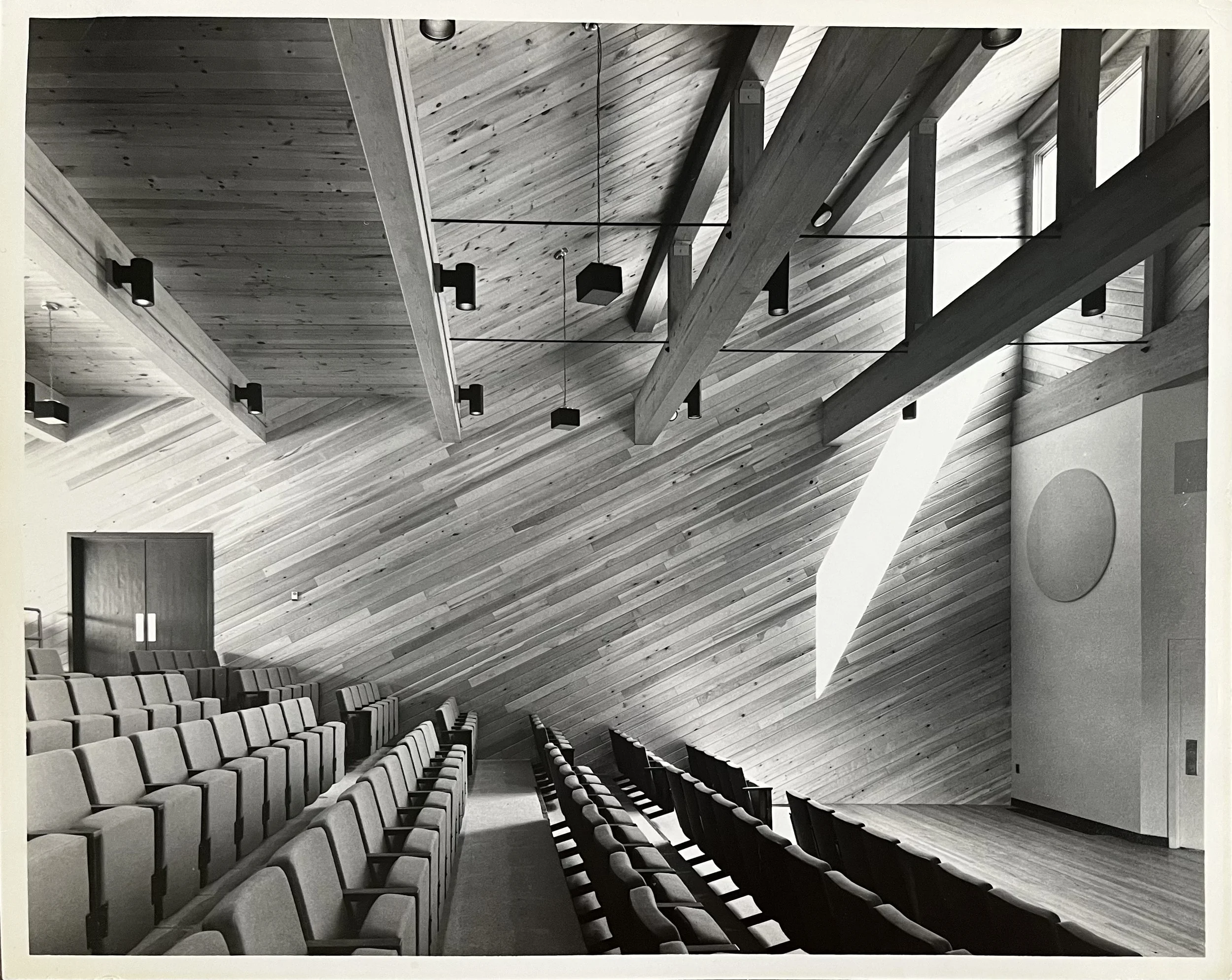 Interior view of an auditorium or lecture hall with rows of cushioned seats, wooden walls, and ceiling with exposed beams and hanging lights.