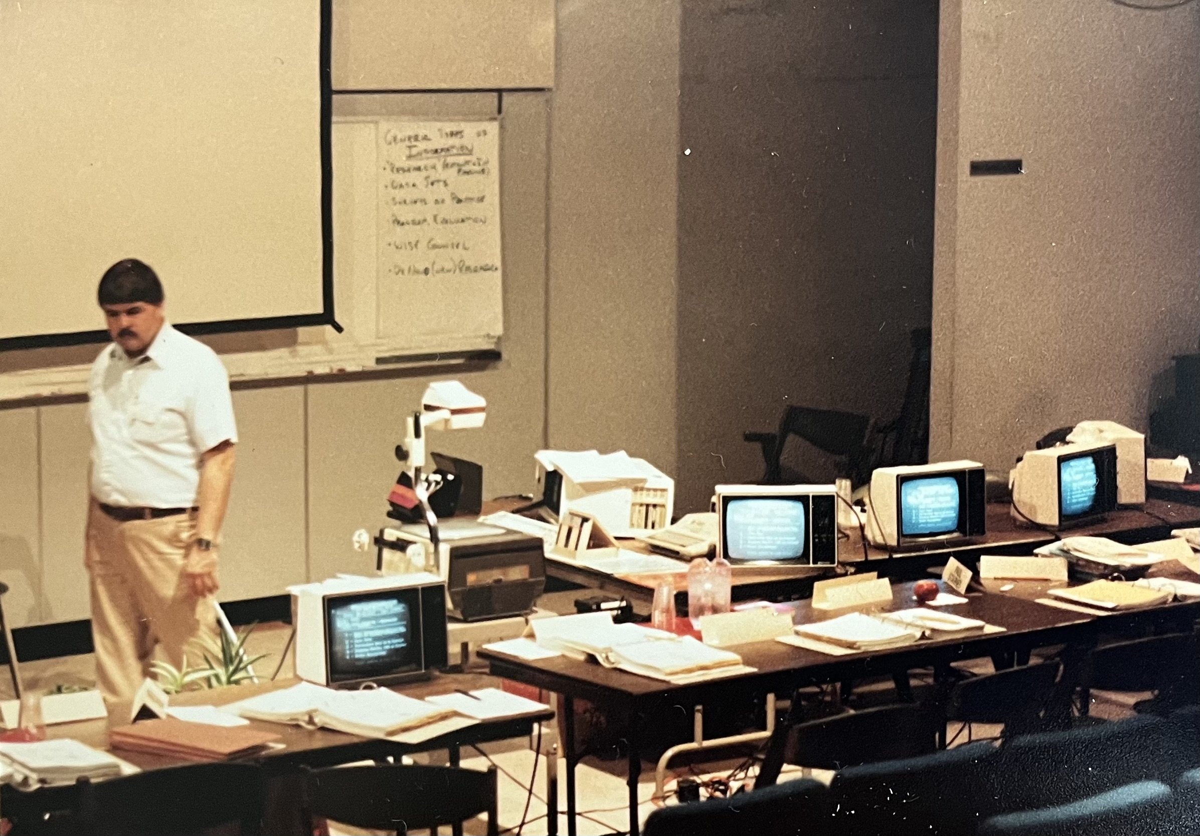 A man with a mustache wearing a white shirt and beige pants standing in a classroom or office setting with multiple old computer monitors, paperwork, and a whiteboard in the background.