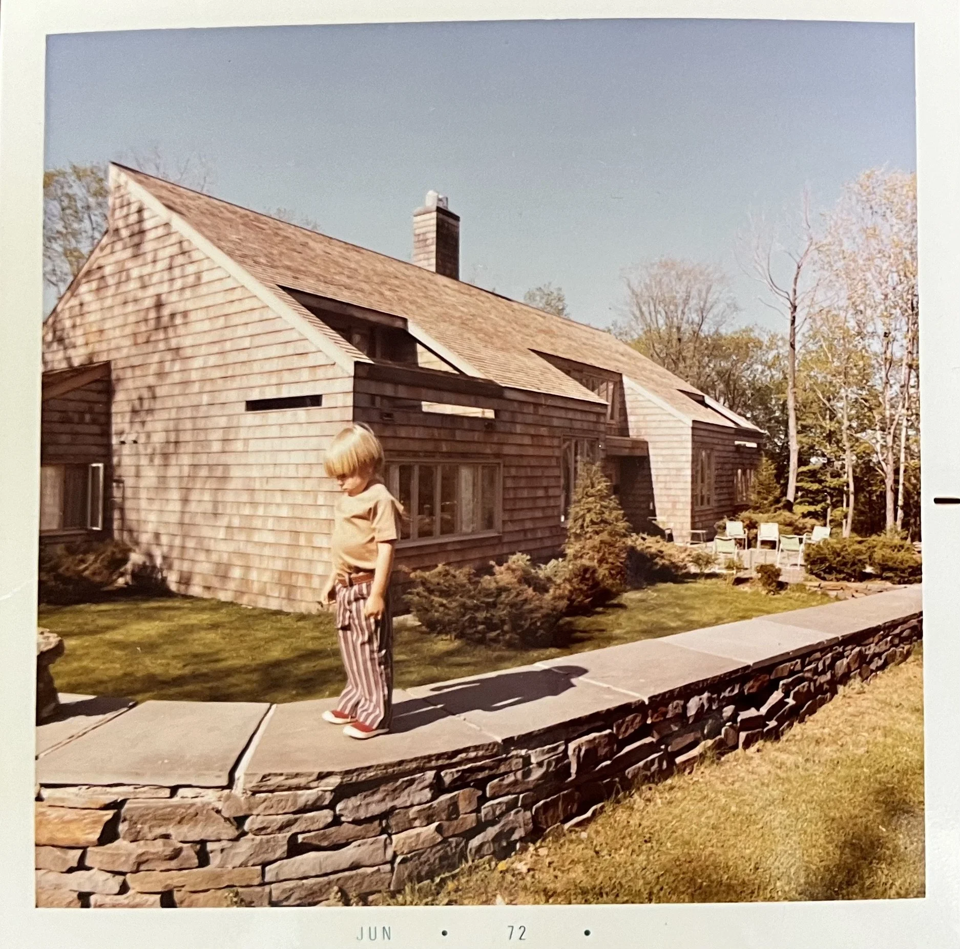 A young boy with blonde hair wearing a beige shirt and striped pants stands on a stone wall in front of a wood-shingled house with large windows, surrounded by trees and a manicured lawn.