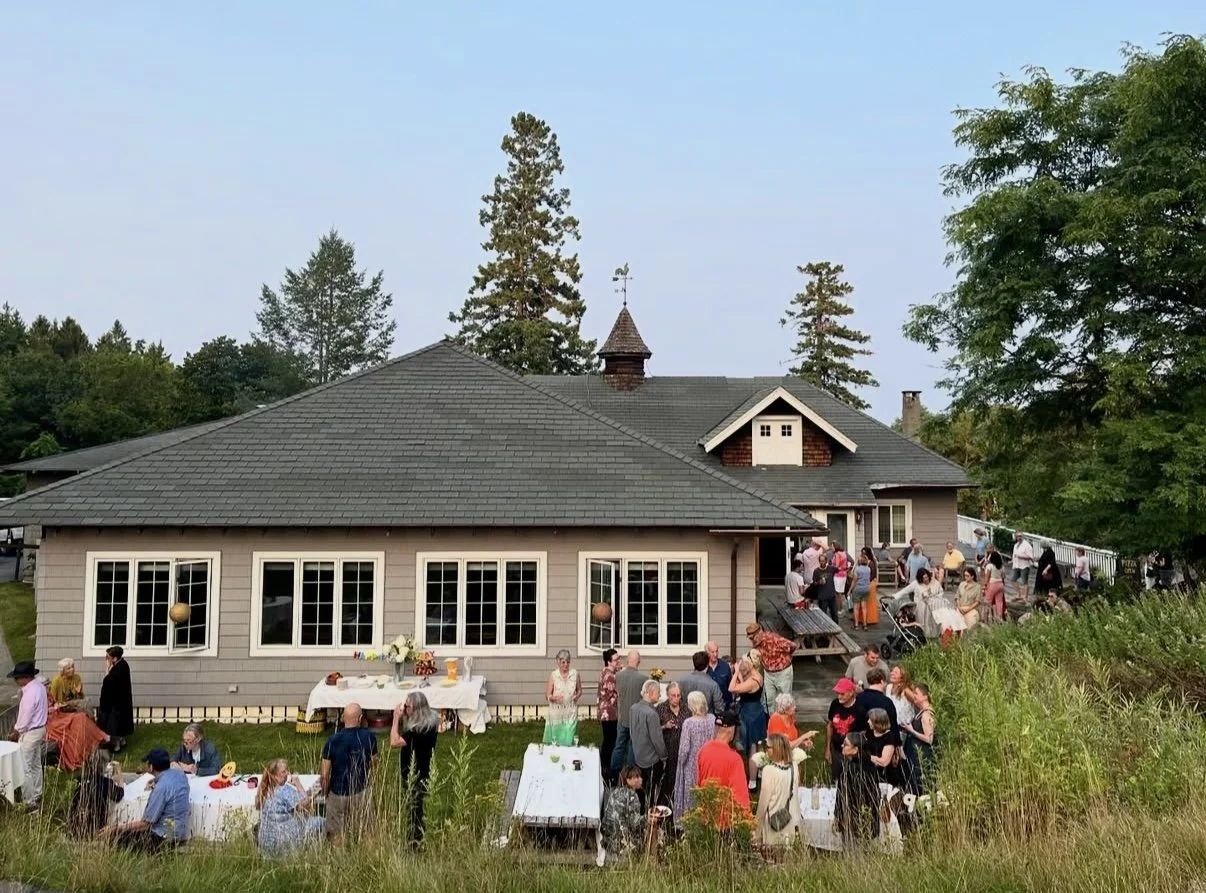Outdoor gathering at a house with people socializing, sitting at tables, and standing on the deck during daytime.