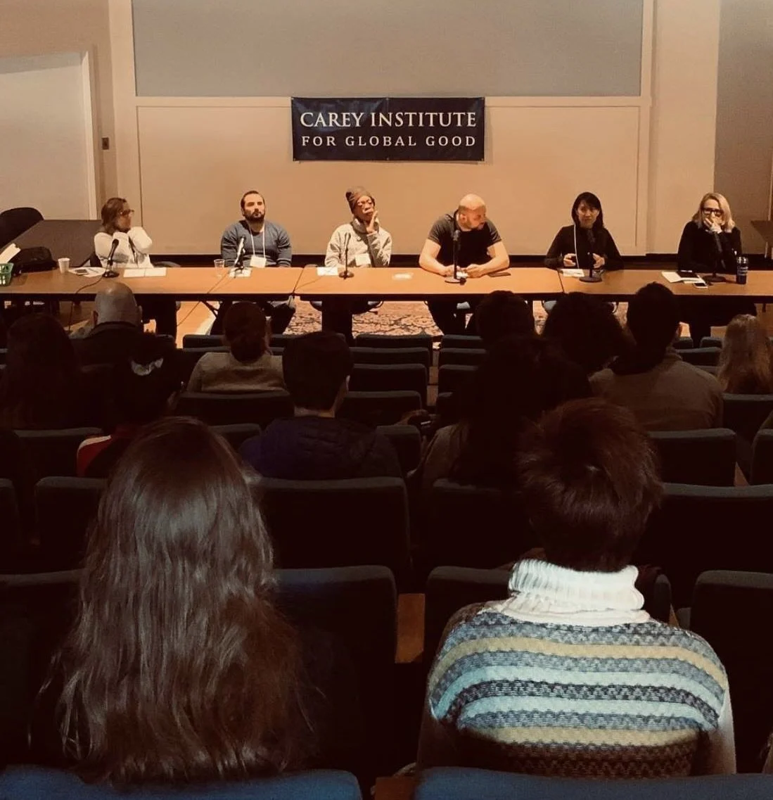 Panel discussion at the Carey Institute, with six people seated at a long table on stage, and an audience watching. A banner reads 'Carey Institute for Global Good'.