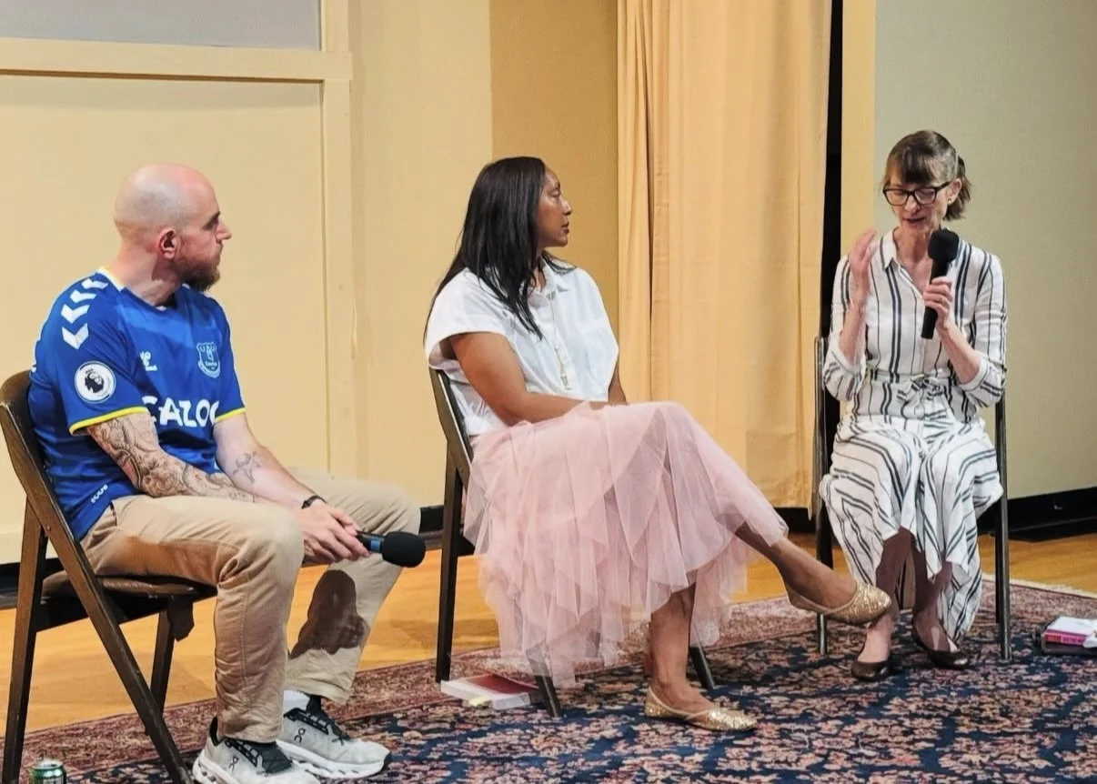 Three people sitting and talking during a panel discussion or interview. One man in a blue sports jersey with tattoos, a woman in a pink tulle skirt and white blouse, and another woman in a striped dress holding a microphone.