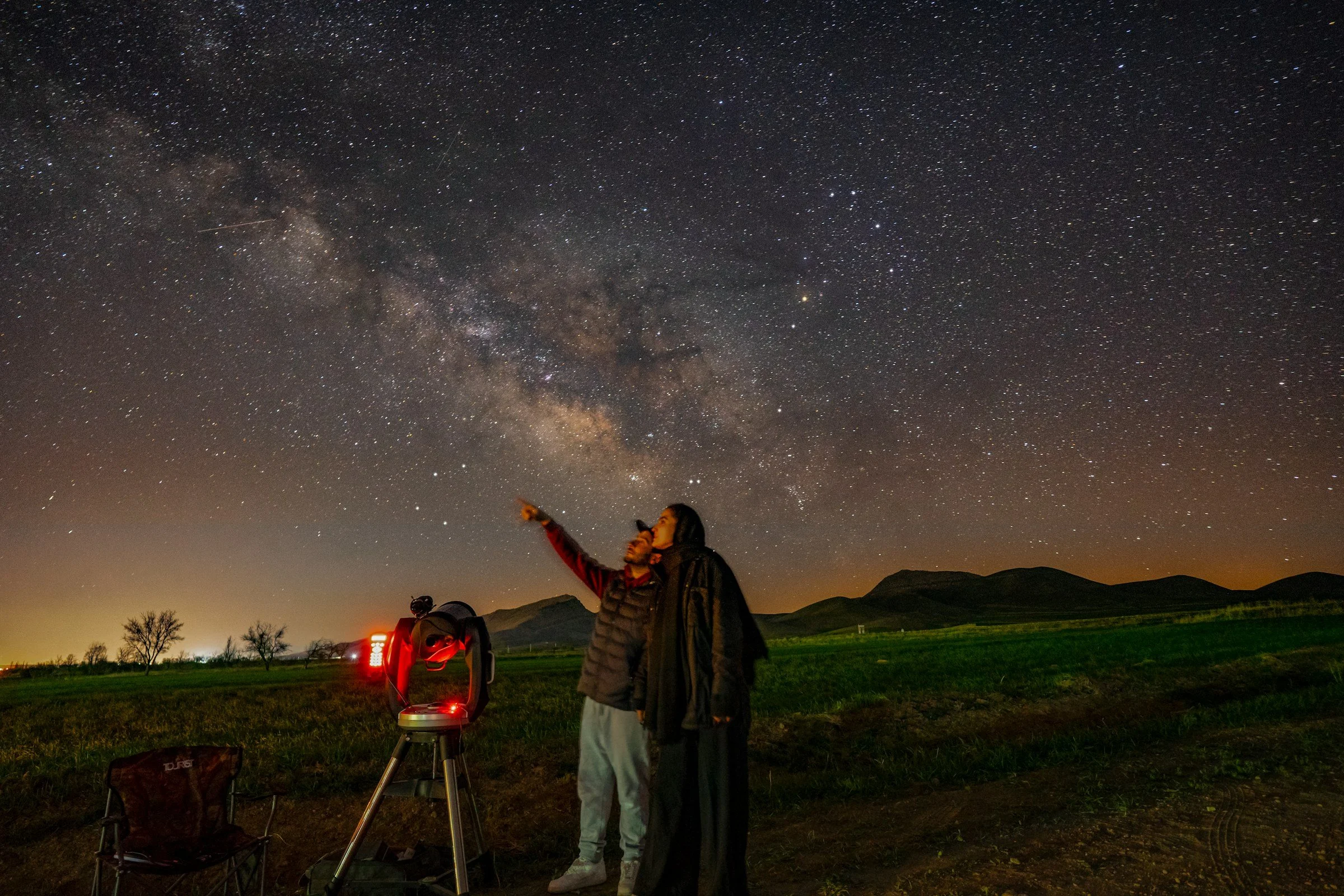 Two people observing the starry night sky with a telescope outdoors. One person is pointing toward the stars, and the Milky Way galaxy is visible in the background.