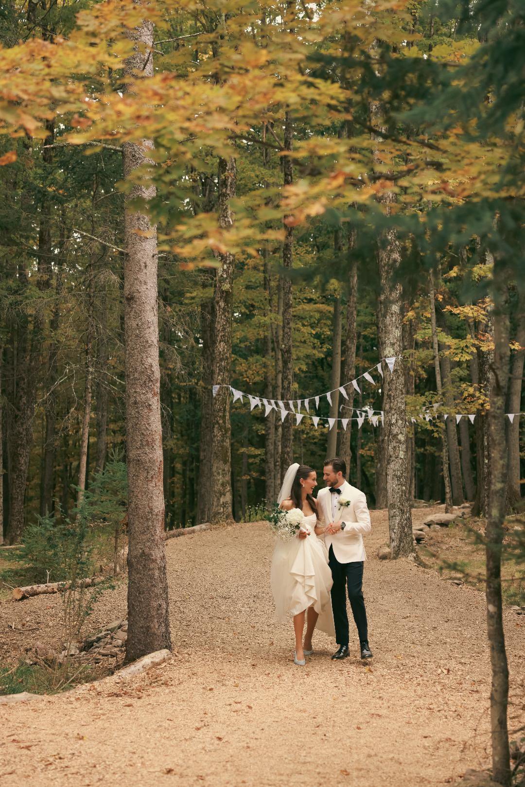 Bride and groom walking together in a forest with autumn foliage, holding hands and smiling, with white bunting hanging between trees above them.