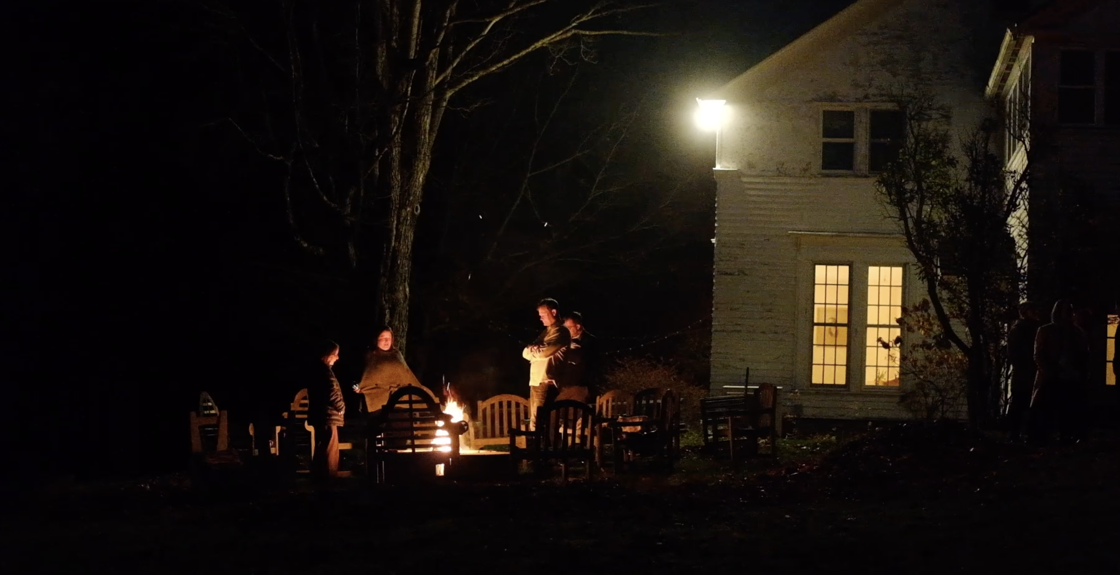 People gathered around a fire pit outside at night, with a multi-story house in the background illuminated by a bright exterior light, and trees framing the scene.