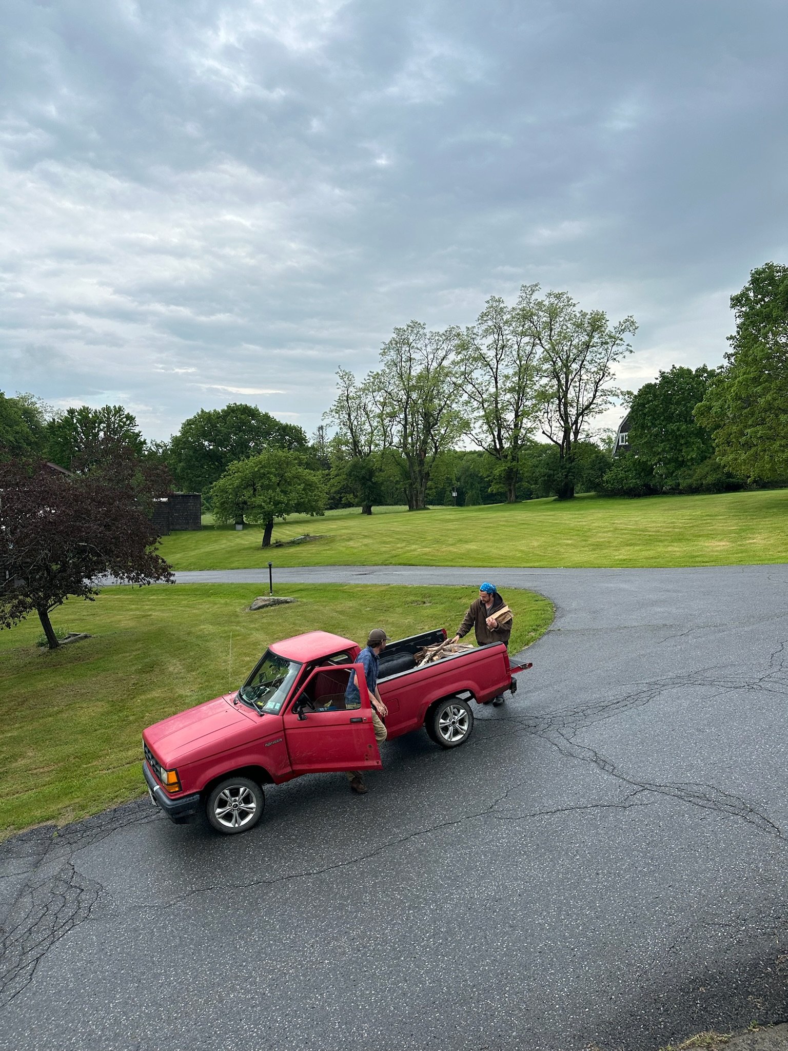 Two men unloading wood from a red pickup truck parked on a driveway in a grassy, tree-filled area with cloudy skies overhead.