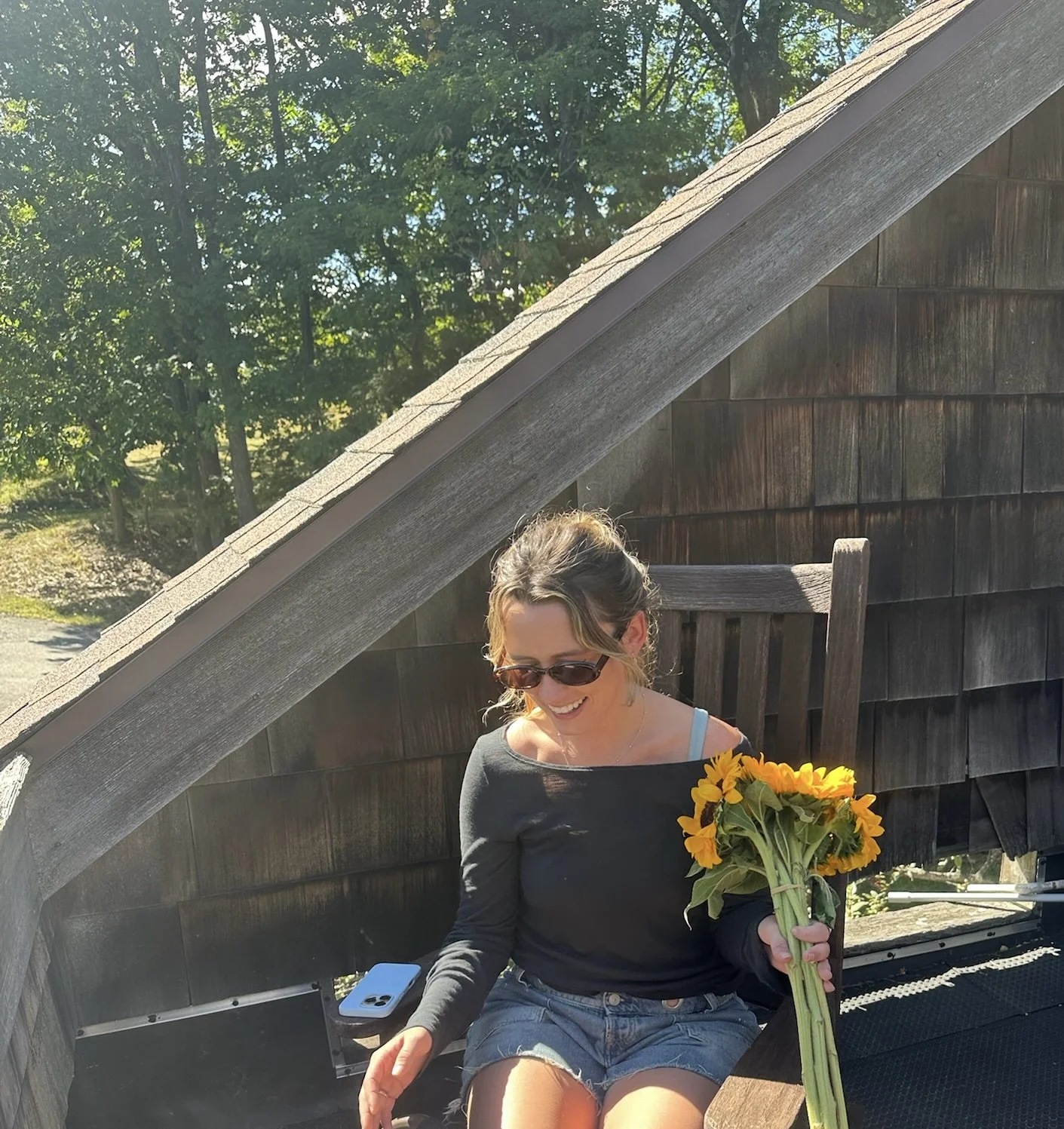 A young woman sitting outside on a wooden chair, holding a bouquet of sunflowers, wearing sunglasses, a black long-sleeve shirt, and denim shorts, smiling and looking down, with a wooden wall and trees in the background.