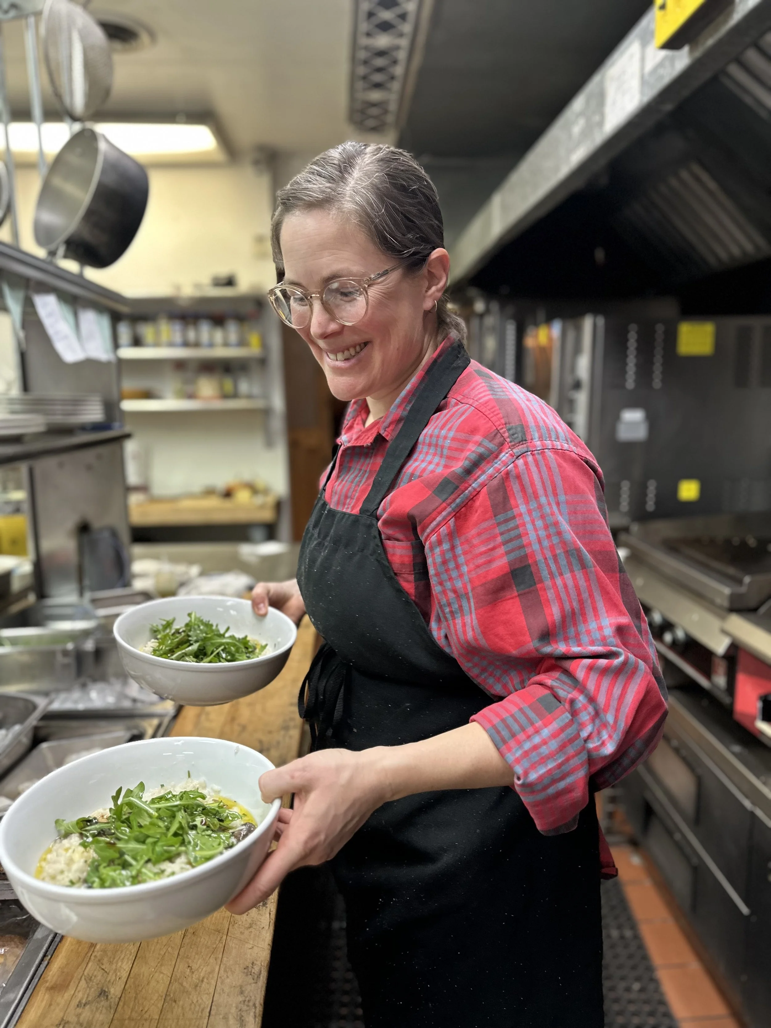 A woman with glasses and a red plaid shirt in a kitchen, holding a white bowl of salad, with more bowls of salad on a wooden counter.