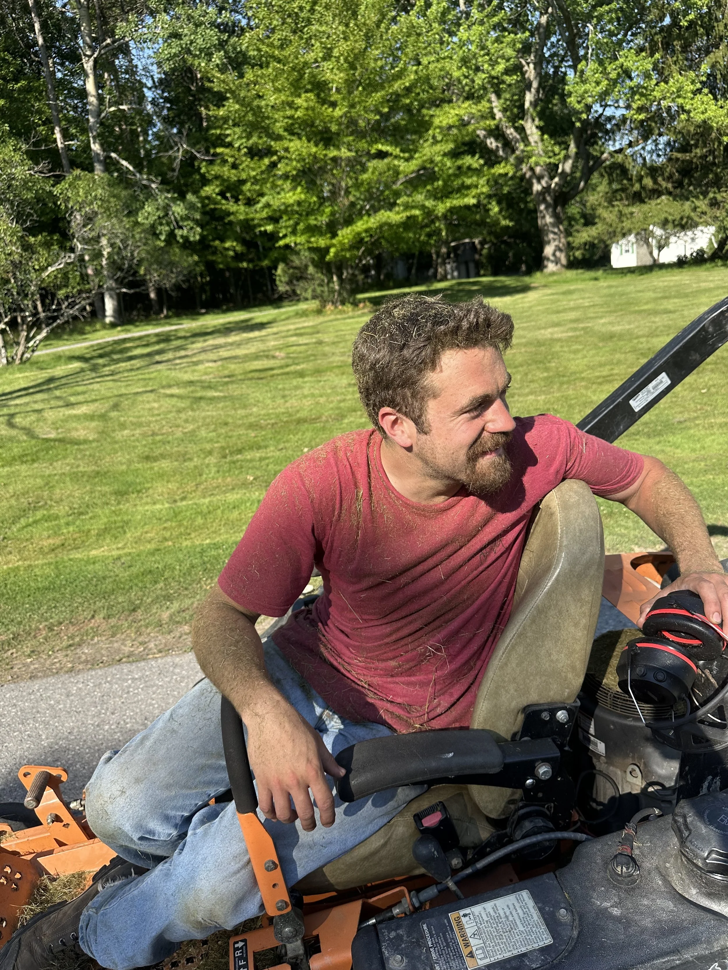 A man with brown hair and beard in a red shirt and jeans, sitting on a riding lawn mower in a grassy yard with trees in the background.