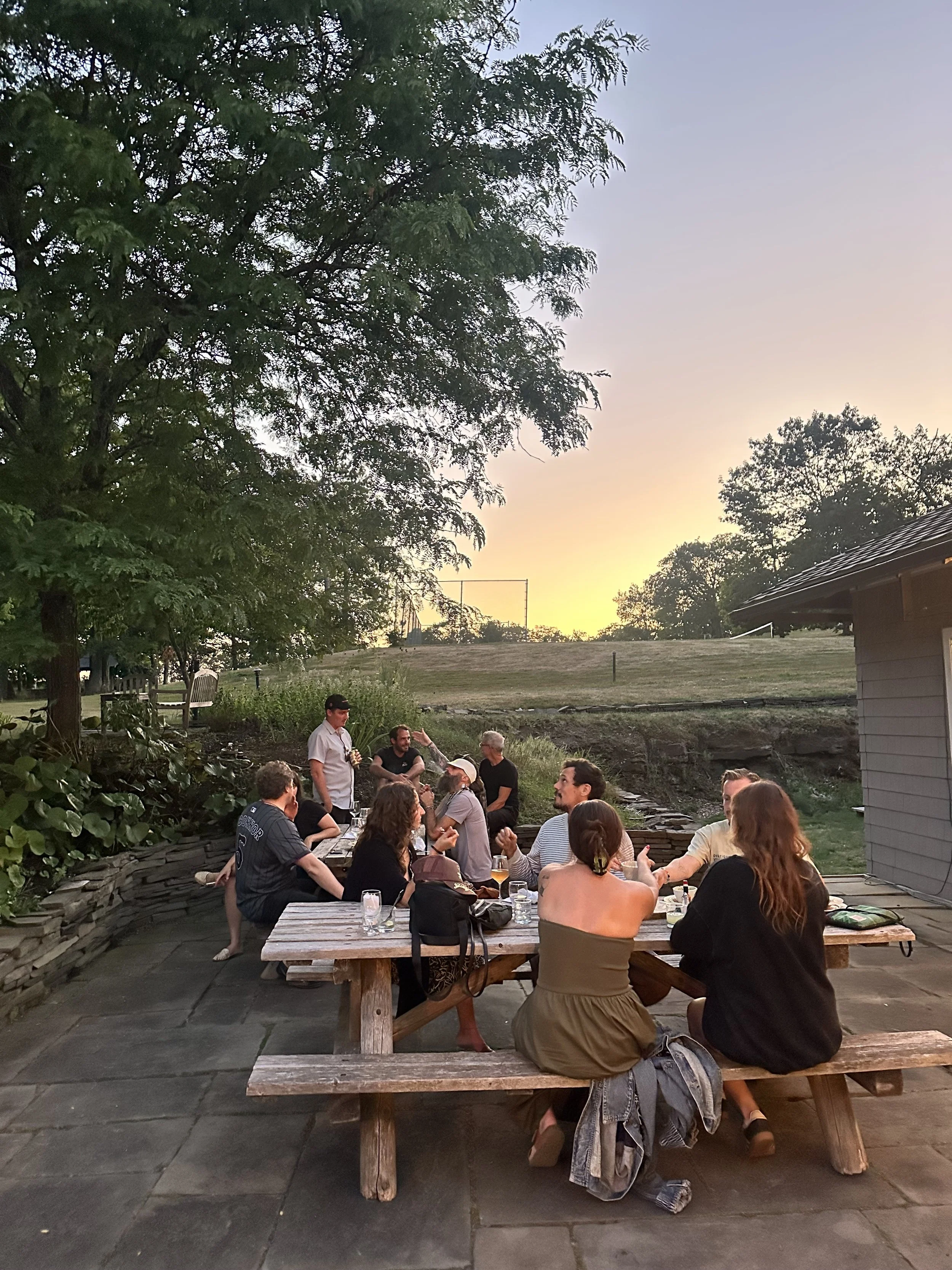 A group of people sitting around a picnic table outdoors during sunset, with trees and a building in the background.