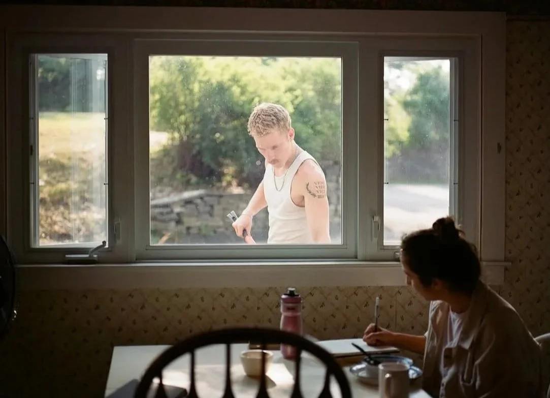 A woman sitting at a dining table inside, looking toward a young man outside through a large window. The young man is wearing a sleeveless shirt, holding a phone, and appears to be outdoors in a natural setting with trees and greenery.