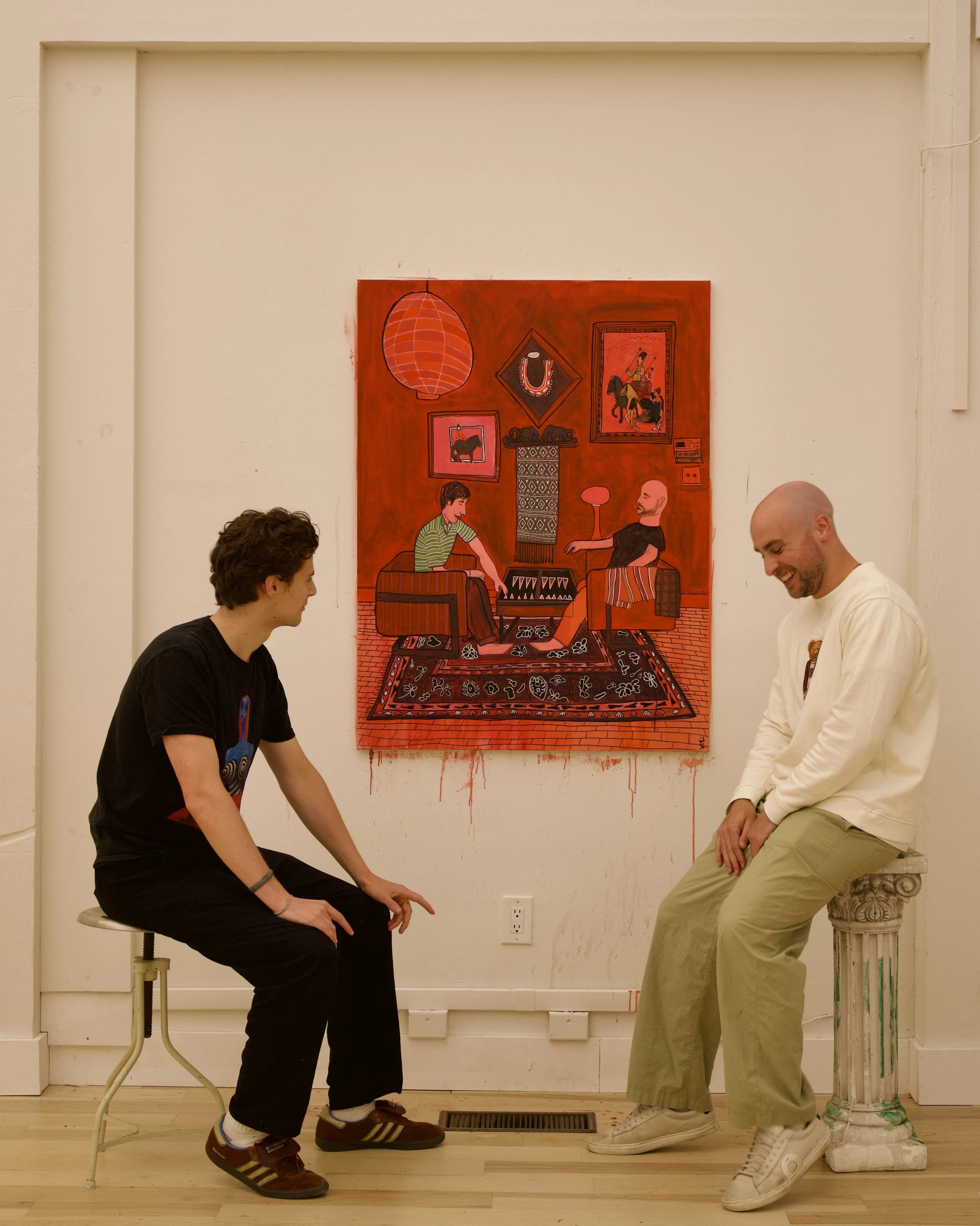 Two men engaging in conversation in front of a red artwork on a white wall. One man, wearing a black t-shirt and black pants, is sitting on a stool and leaning forward, while the other man, wearing a white sweatshirt and khaki pants, is sitting on a 
