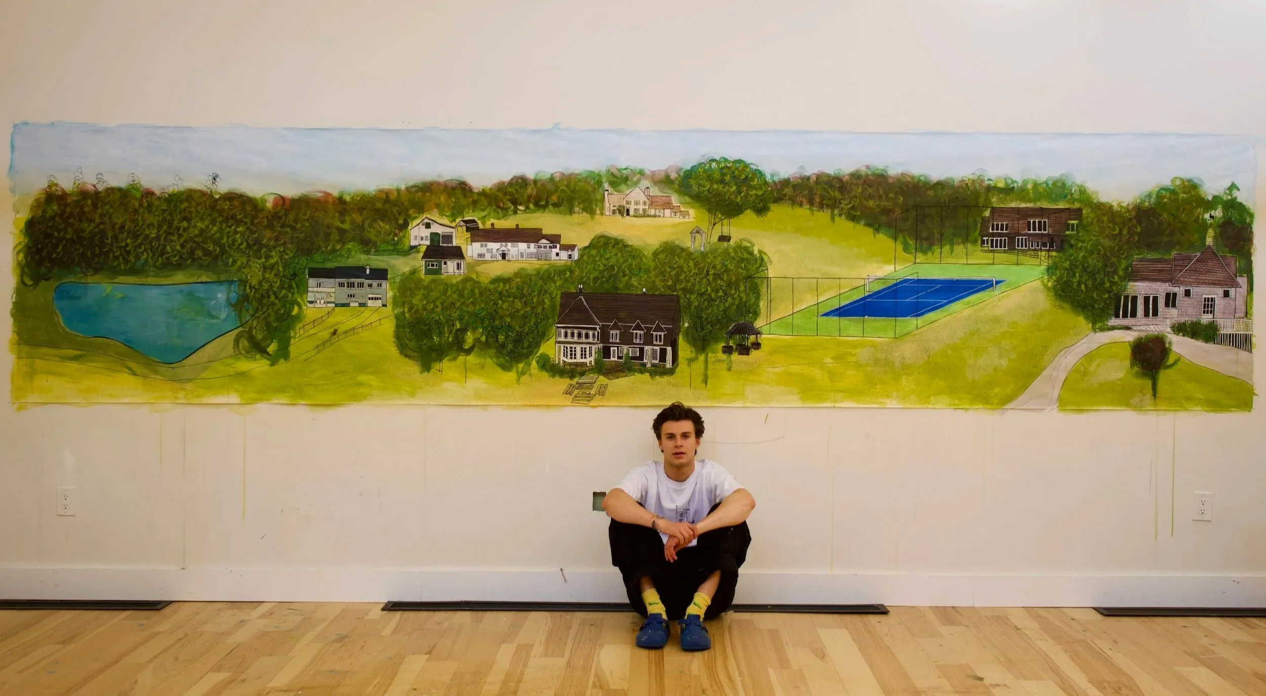 Young man sitting cross-legged in front of a large hand-painted landscape mural on a wall, featuring houses, trees, a pond, and a tennis court.