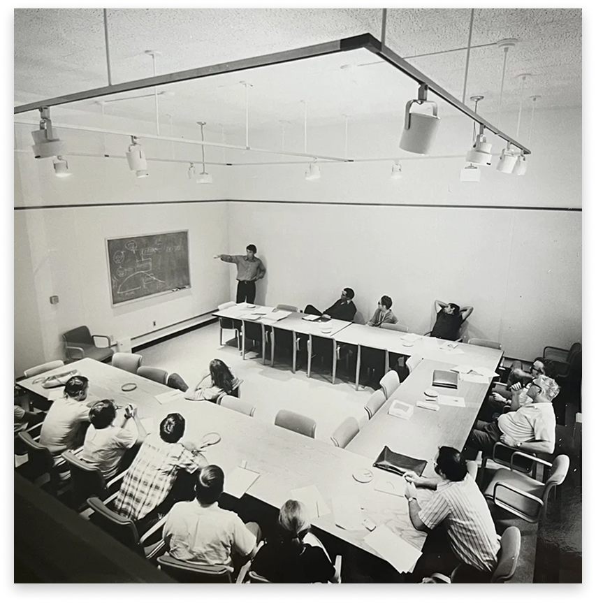 A black-and-white photo of a classroom with several students seated around a U-shaped table, and a teacher at the front pointing at a blackboard with diagrams or drawings.