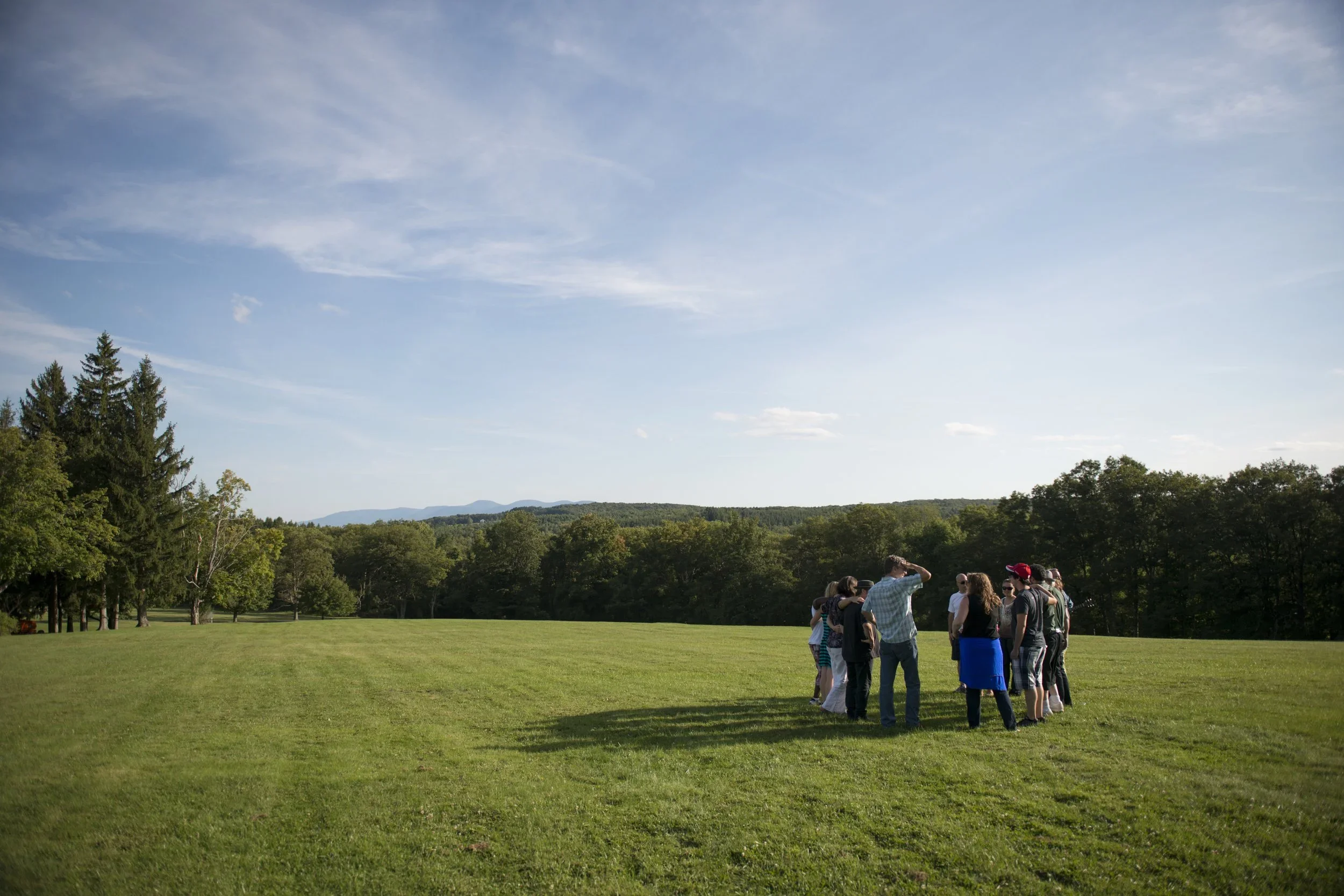 A group of people gathered outdoors on a grassy field with trees, mountains, and a partly cloudy sky in the background.