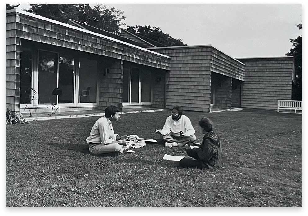 Three people sitting on the grass in front of a modern-style house, engaged in conversation and reading, with a bench and large windows in the background.