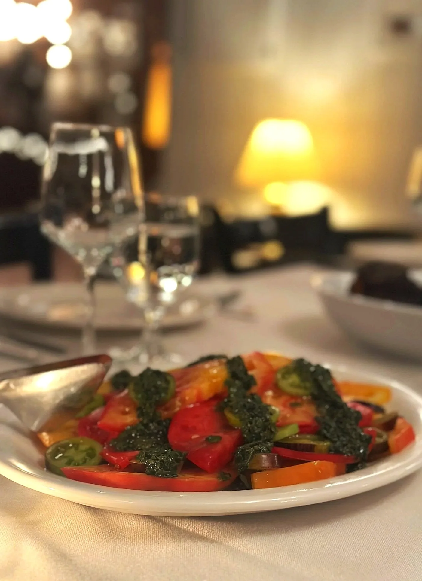 A restaurant table with a plate of colorful roasted vegetables topped with green herb sauce, a wine glass, and a blurred background with warm lighting.
