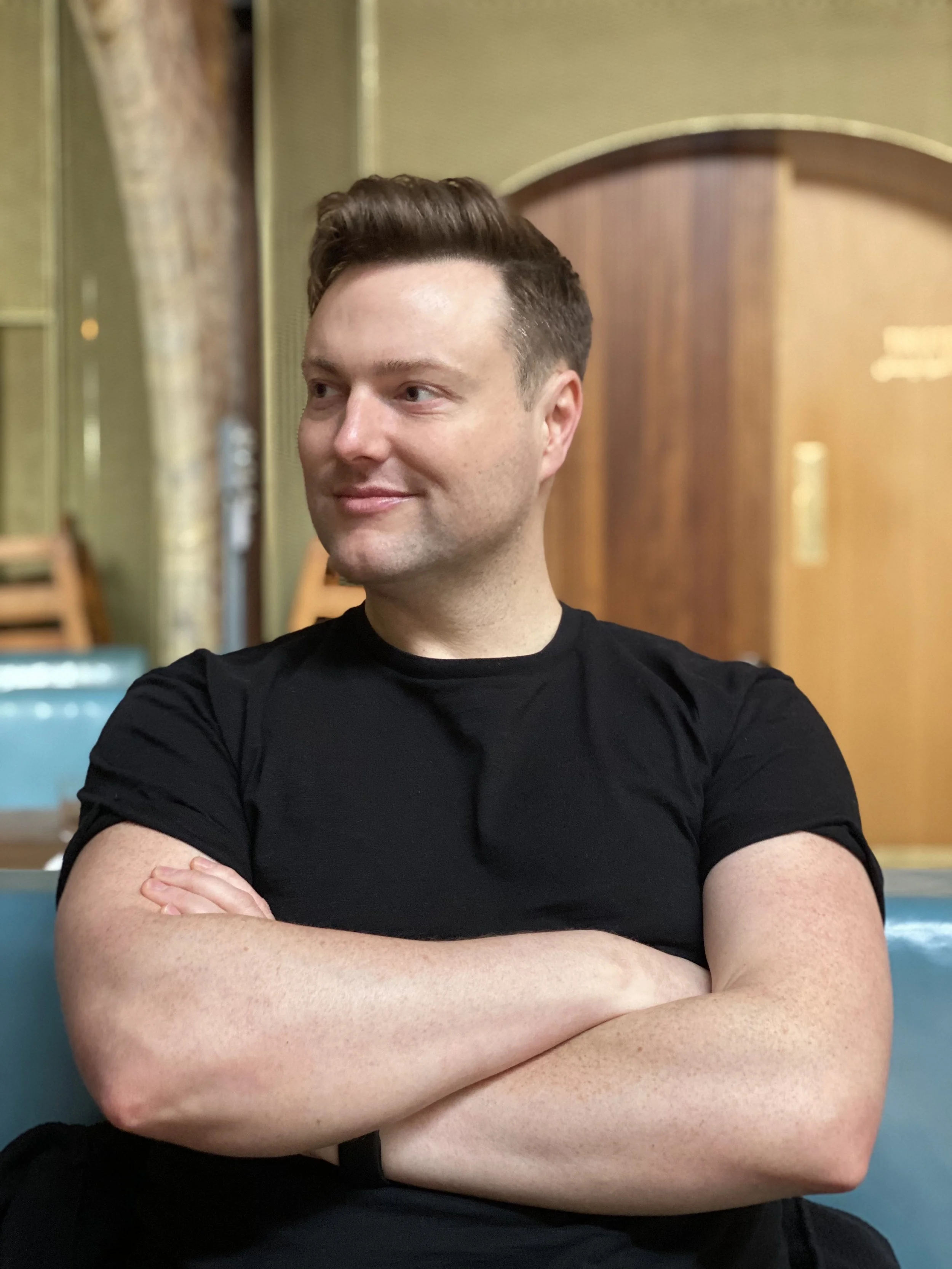 A man with short, styled brown hair sitting with his arms crossed, wearing a black t-shirt, with a slightly smiling expression, in an indoor setting with wooden and decorative elements in the background.