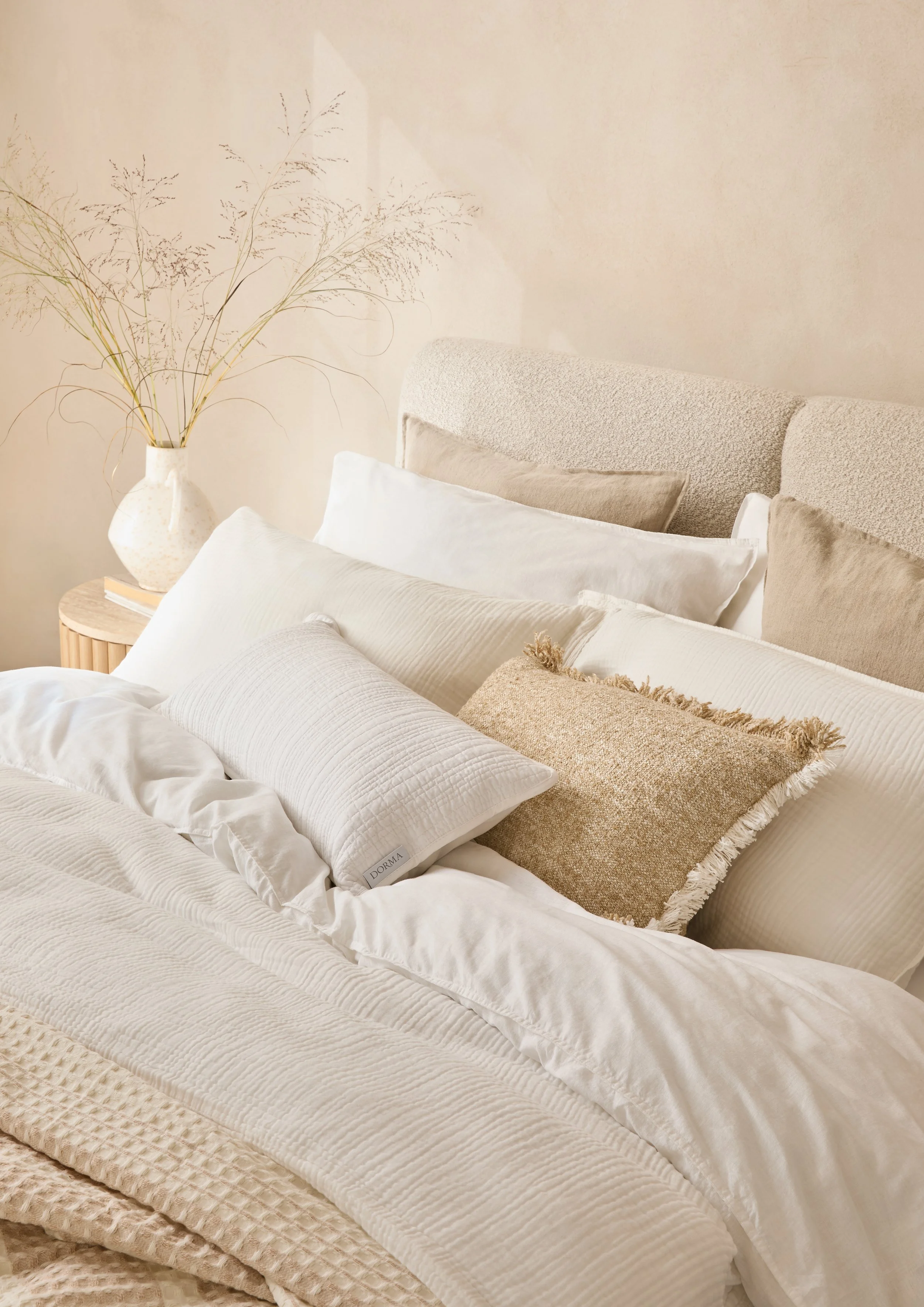 A bed with cream-colored bedding and several pillows, including a textured tan pillow, and a beige headboard, next to a wooden nightstand with a white vase holding dried branches.