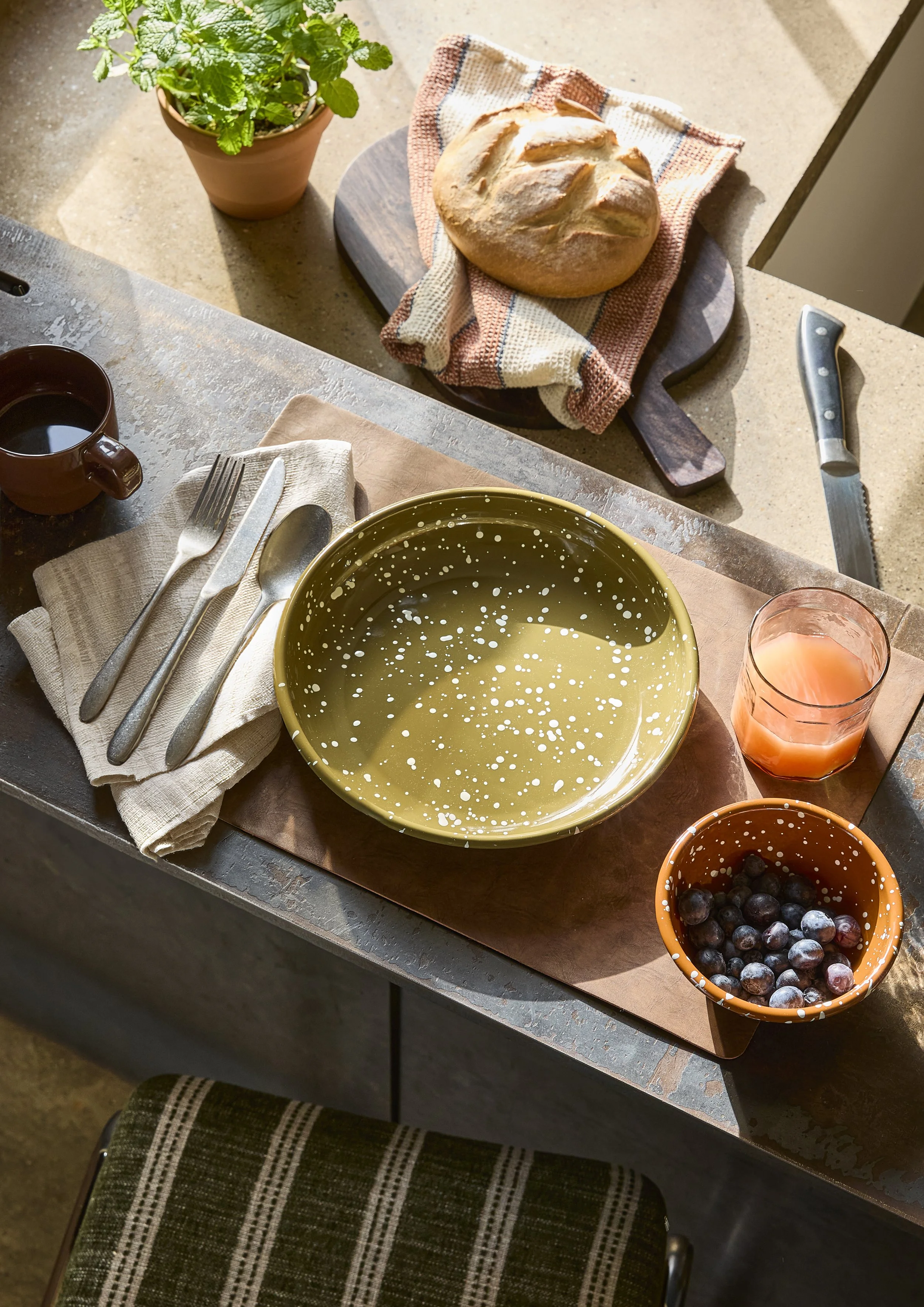 A table with a loaf of bread on a cutting board, a potted mint plant, a yellow and white speckled bowl, a cup of coffee, and a glass of orange juice. There is also a bowl of blueberries and a set of silverware on a cloth napkin.