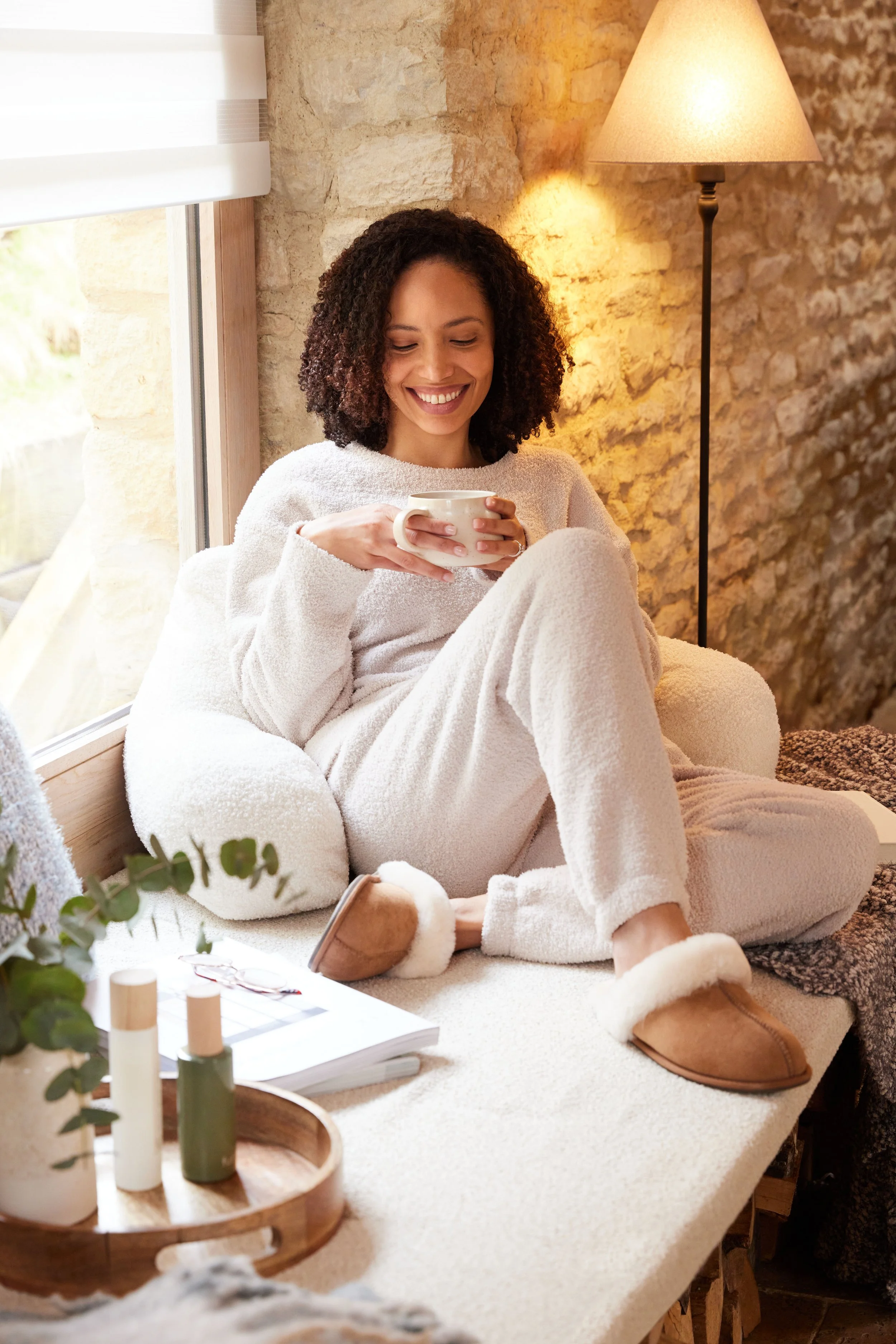 A smiling woman with curly hair sitting on a cozy beige blanket near a window, holding a white mug, with a stone wall and a standing lamp in the background, and decorative items on a table in the foreground.