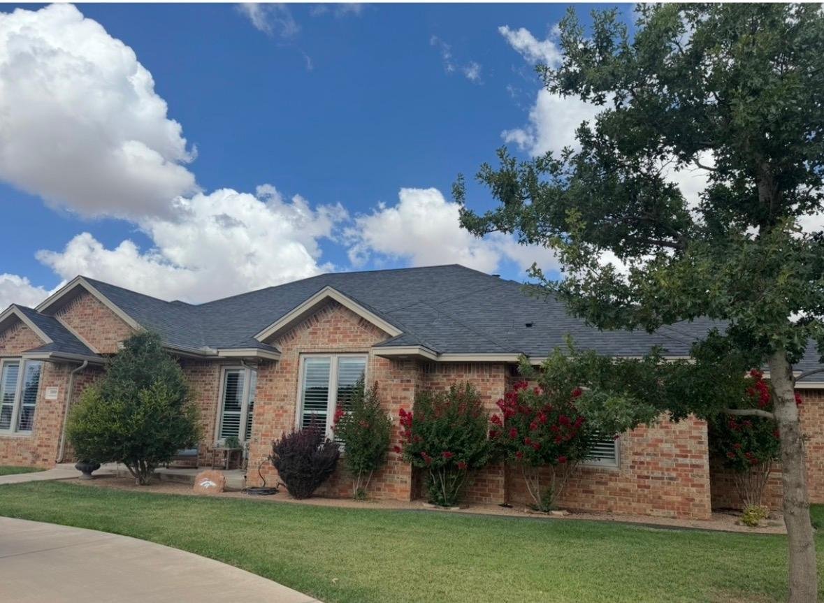 A brick house with a dark gray roof, white window shutters, a small lawn, bushes with red flowers, and a large tree under a blue sky with white clouds.
