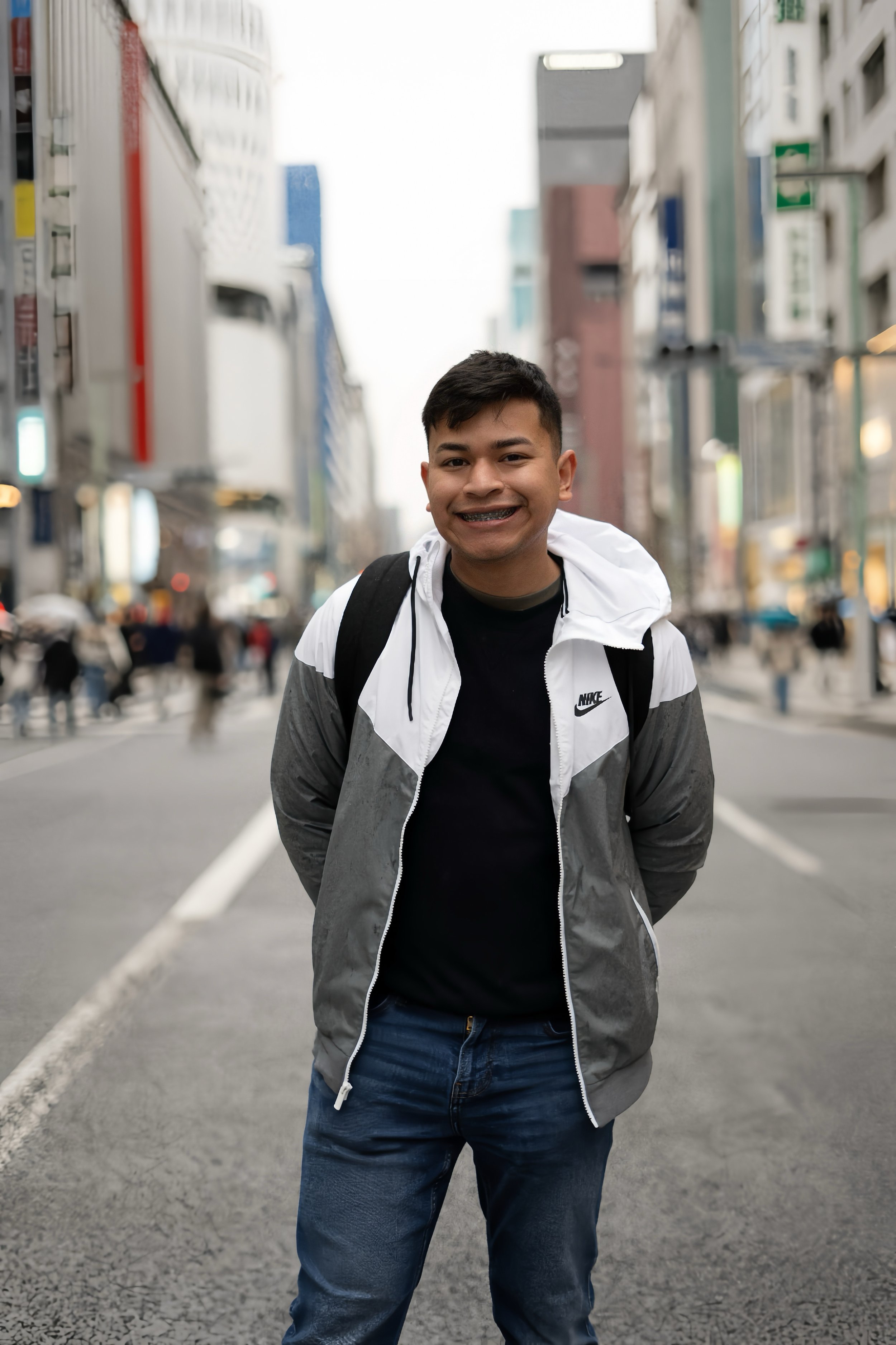 A young man smiling and standing with his hands in his pockets on a busy city street, wearing a white and gray jacket, black shirt, and jeans.