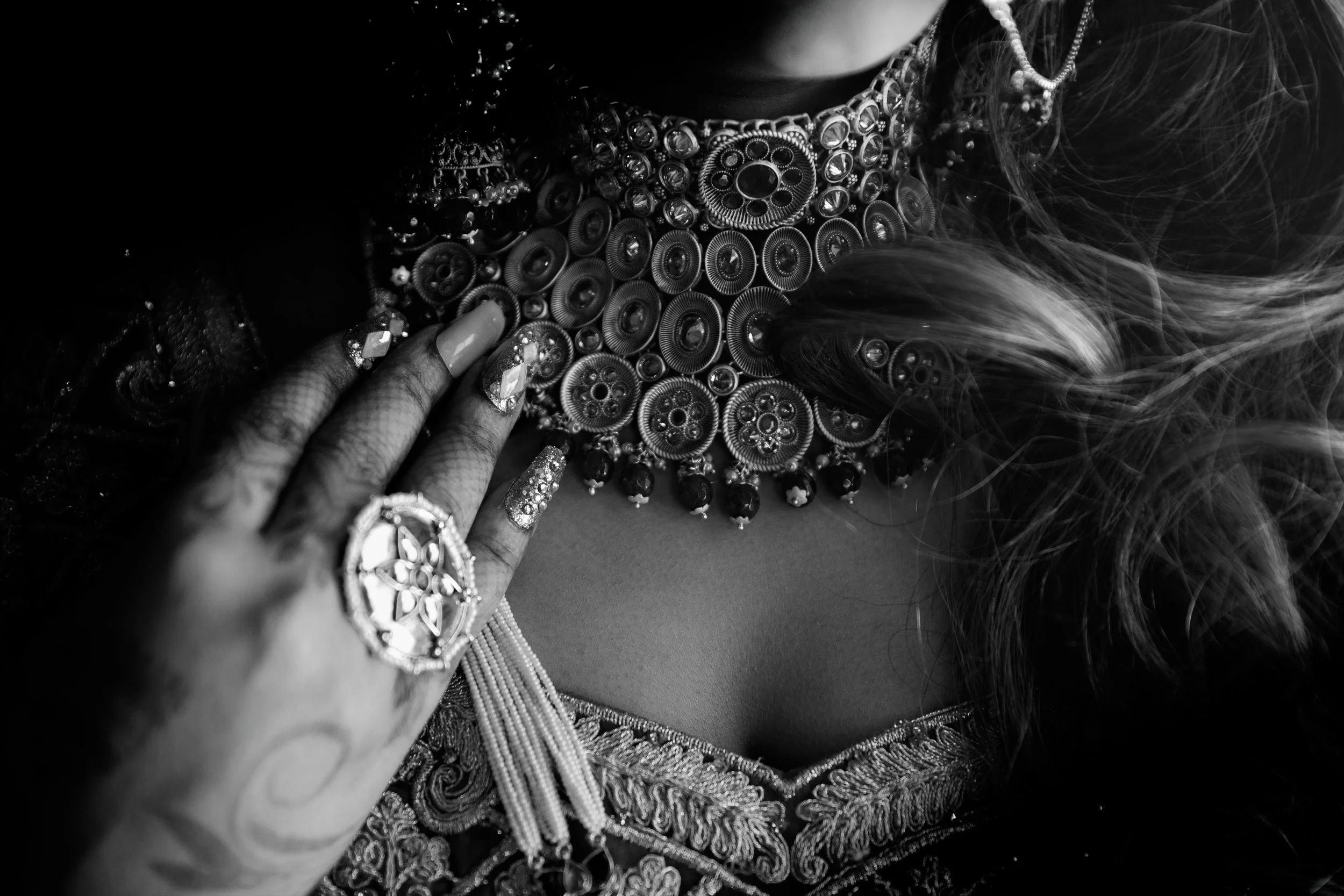 Close-up of a woman's neck and hand showcasing elaborate jewelry including a large statement necklace, rings, and nails decorated with rhinestones, black and white image.