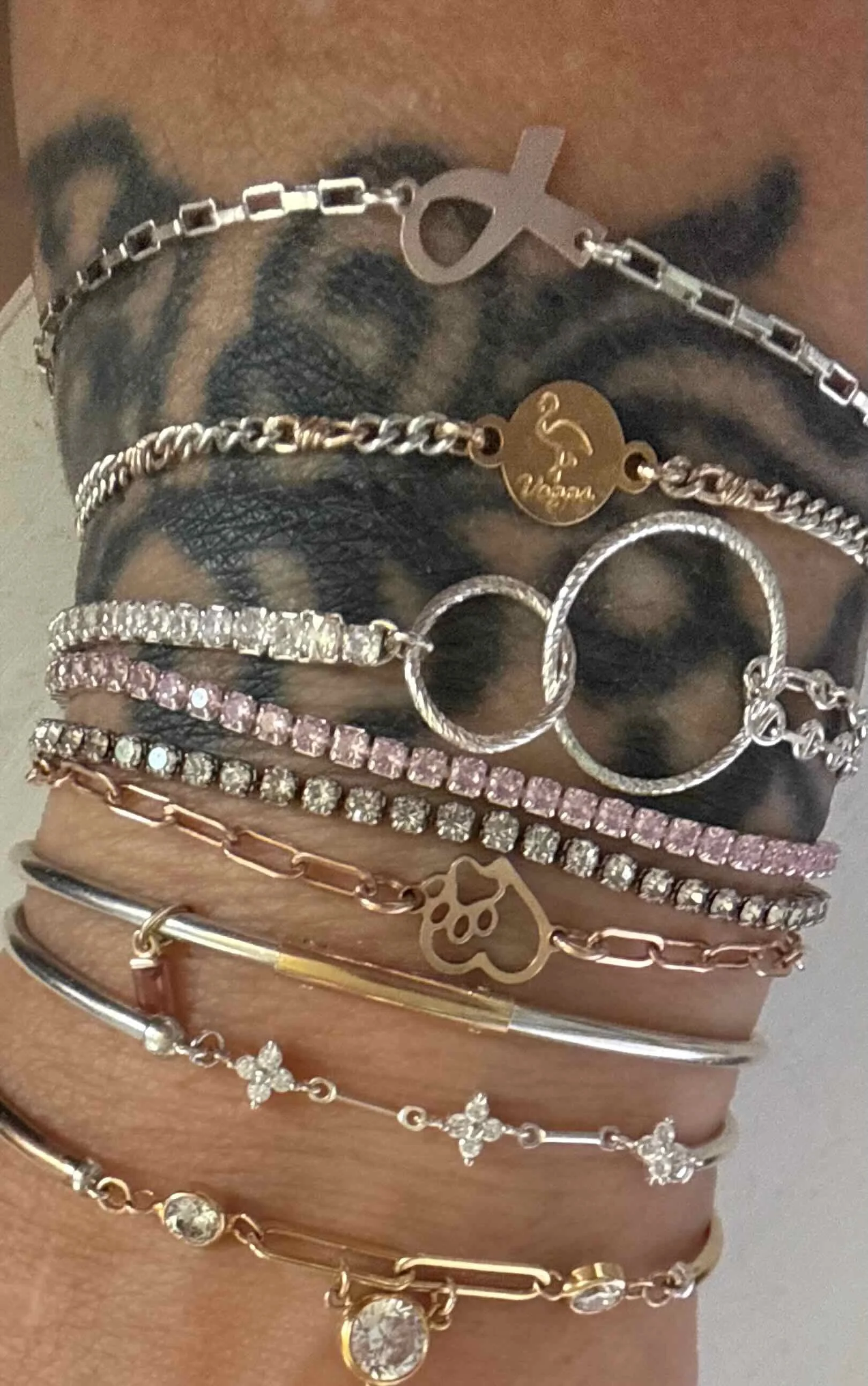 Close-up of a person's wrist adorned with multiple layered bracelets, including chain bracelets, a beaded bracelet with a heart charm, and a bracelet with a large gemstone, with some wearing rings. Laser engraved charms.