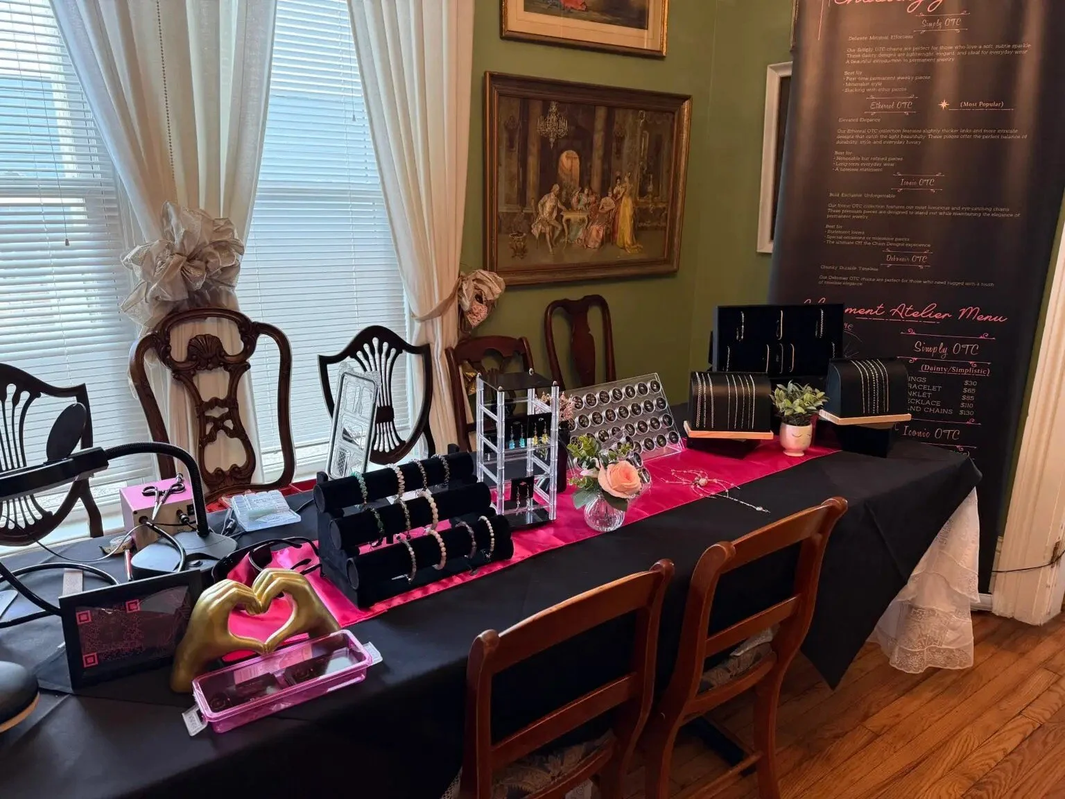 Display of jewelry and accessories on a table with black and pink tablecloths, inside a room with window blinds and framed paintings. Jewelry items include necklaces, bracelets, and earrings arranged on black velvet displays. There is a small potted plant and a pink rose on the table, along with a signboard listing jewelry prices and options.