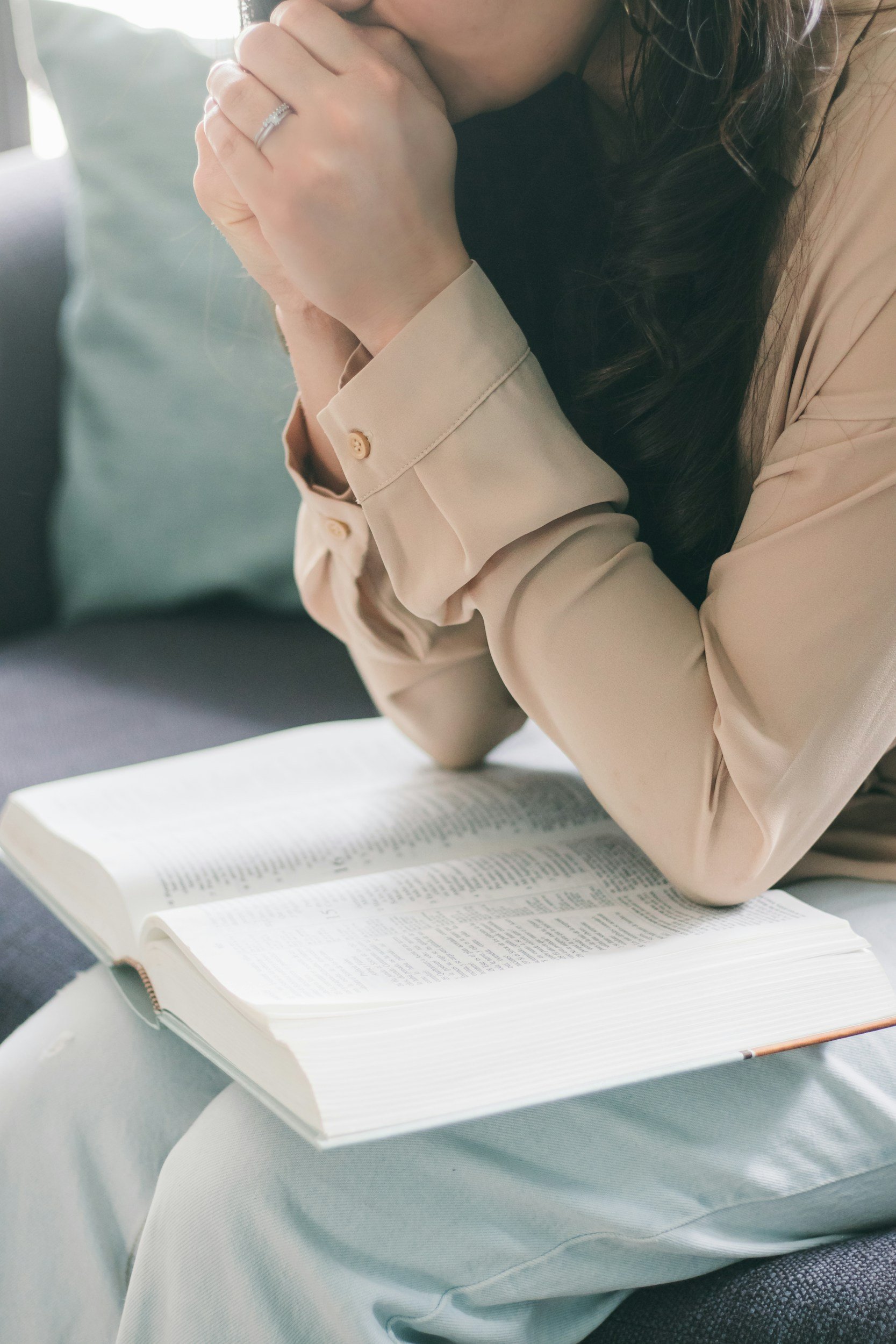 A woman with long dark hair, wearing a beige blouse, sitting with her head resting on her hands, reading a large open book, with a green pillow in the background.