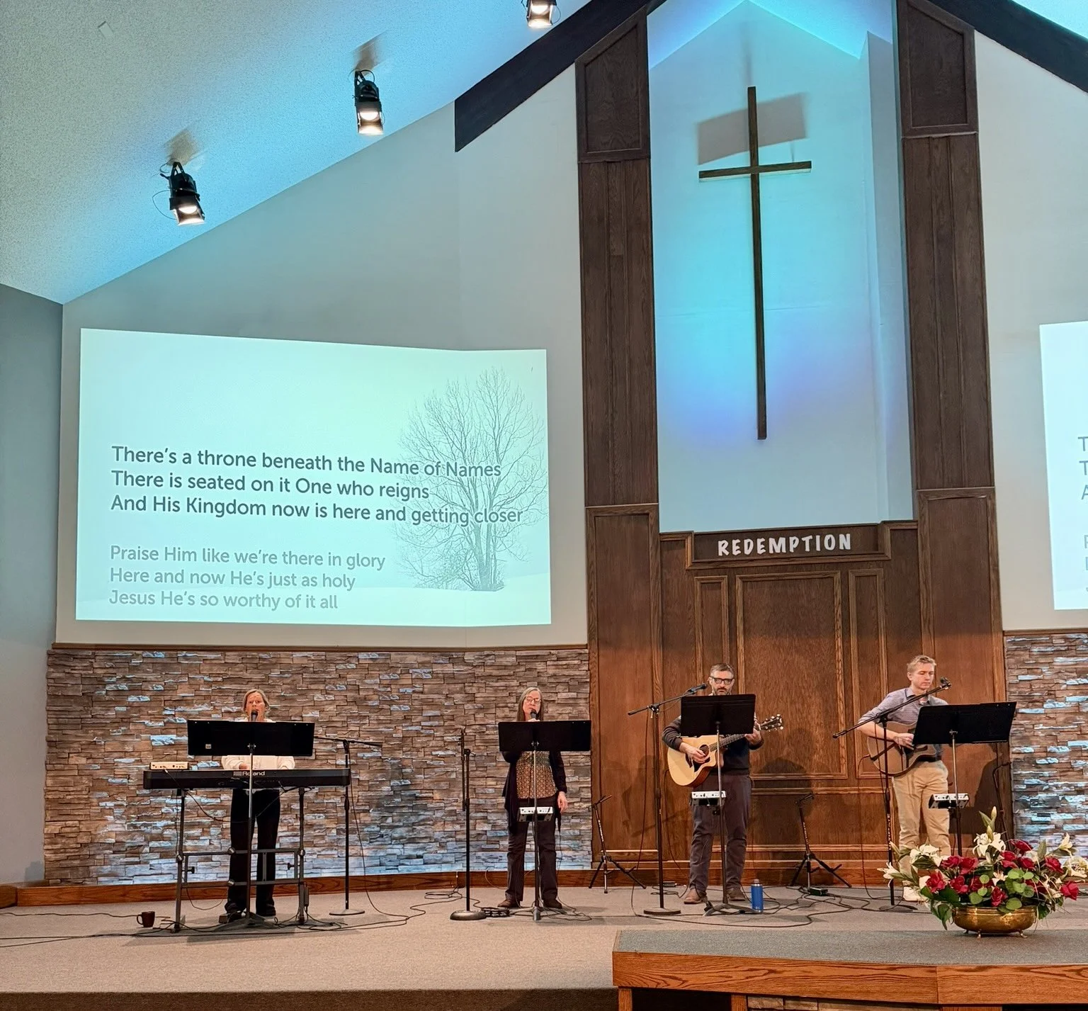 Church choir performing on stage with a large projected hymn lyrics on the wall, a cross with a blue backlight, and a sign reading 'Redemption' above the stage.