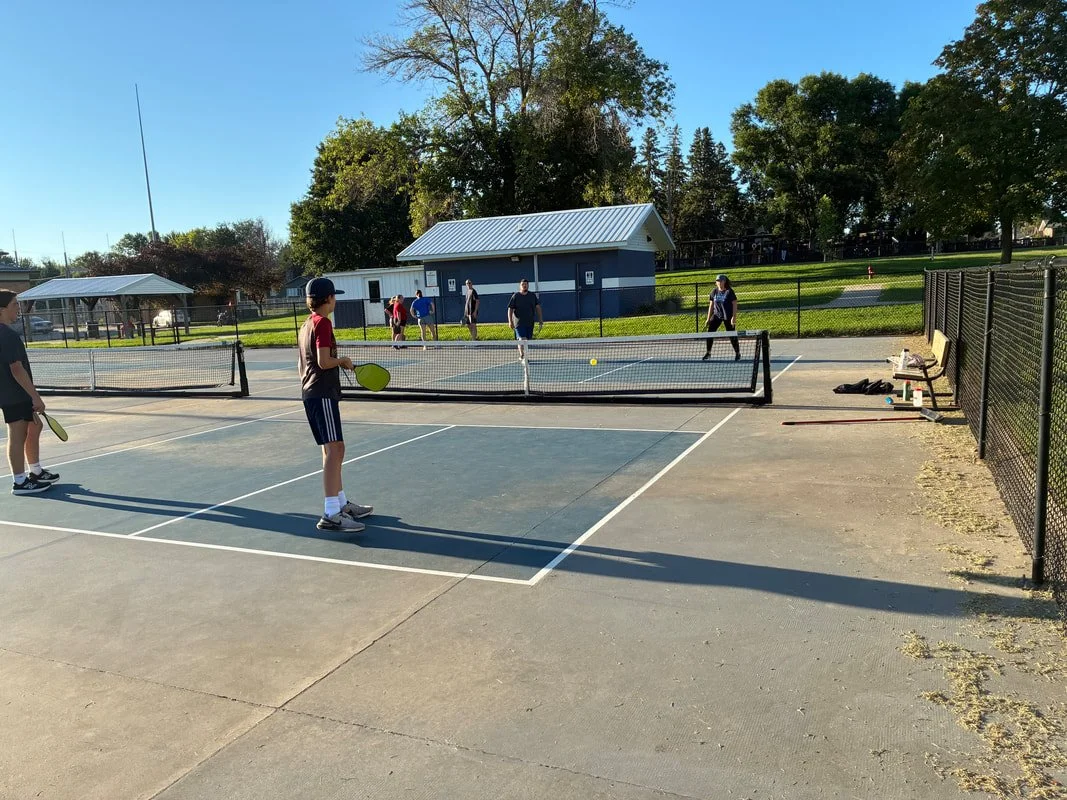 Two children playing pickleball on an outdoor court, with other people in the background and a small building and trees around the court.
