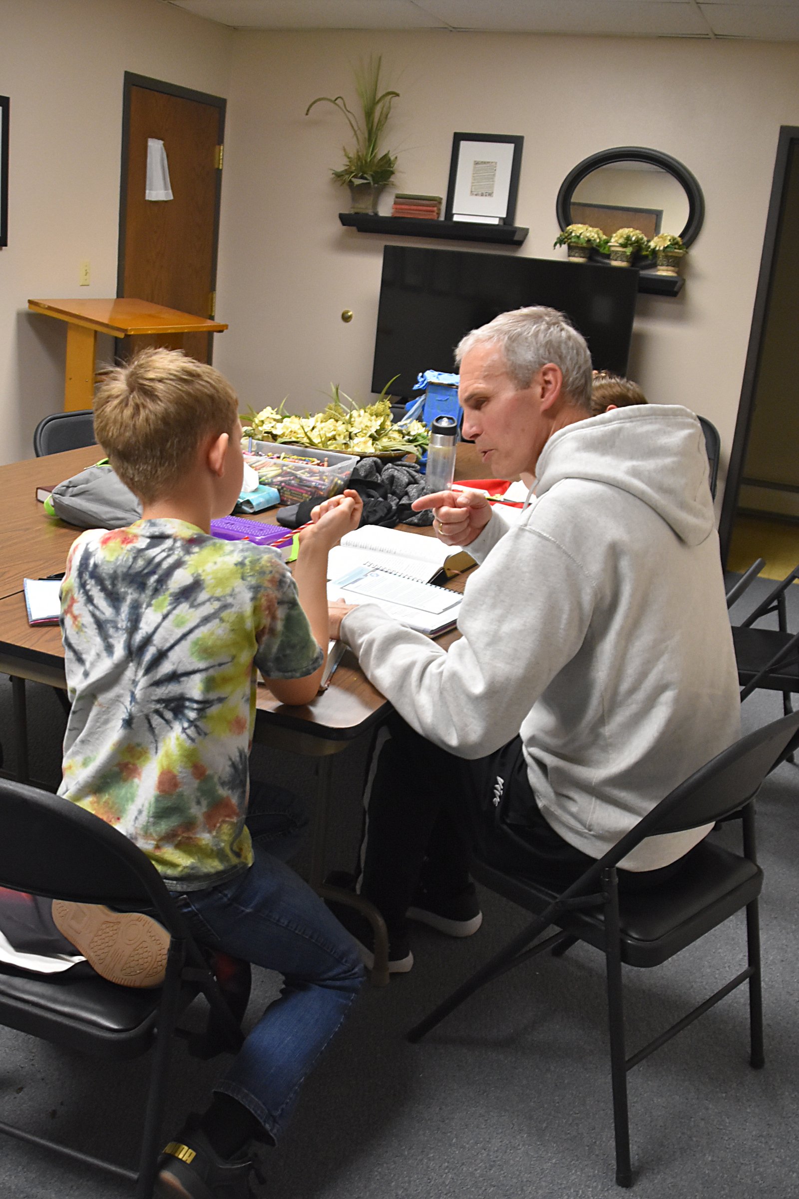 A man with gray hair and wearing a light gray hoodie engaging in conversation with a young boy in a colorful tie-dye shirt, seated at a table in a room decorated with plants, framed artwork, and a large TV.